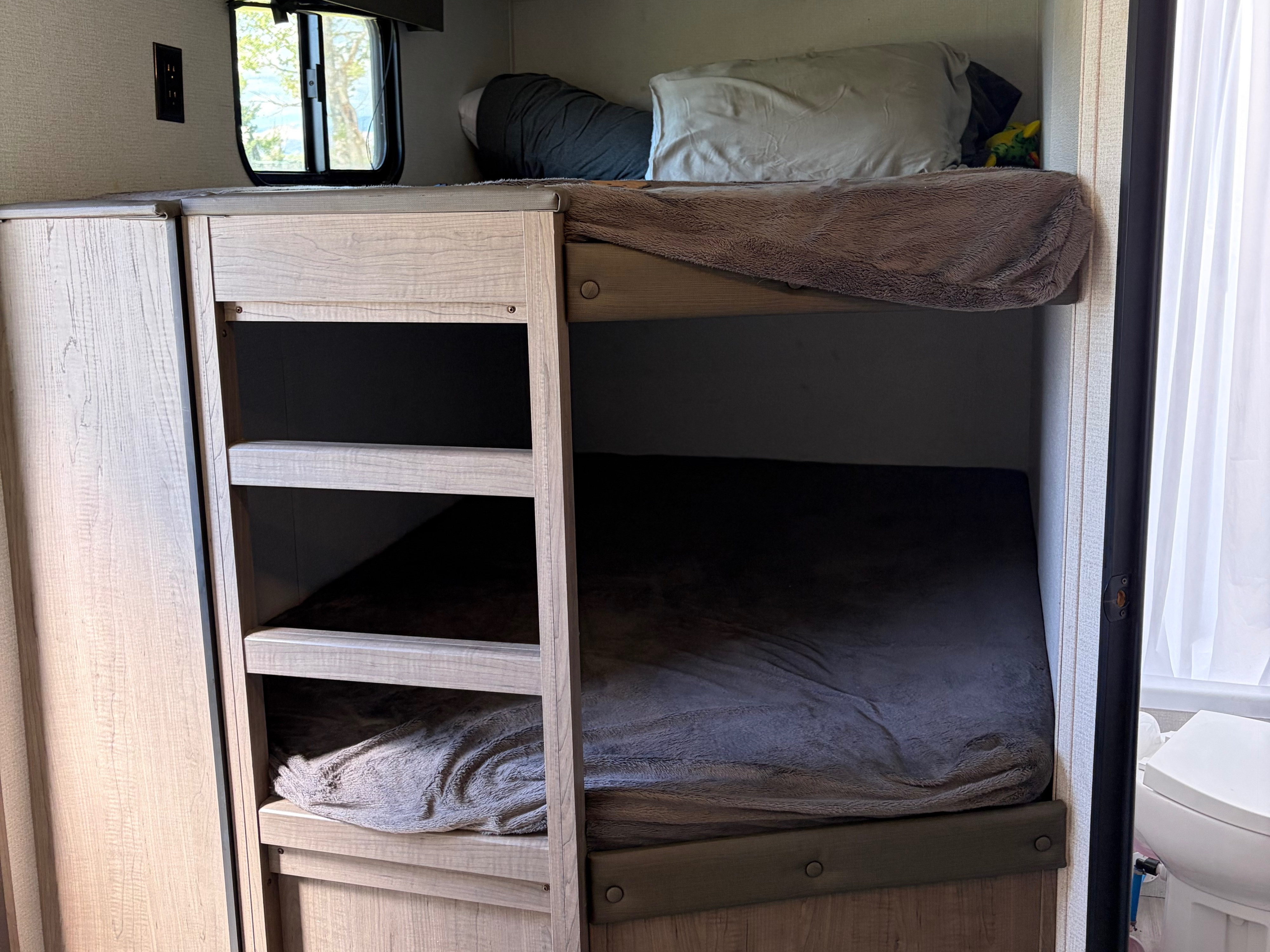 Inside an RV rental: light wood bunk beds with bedding, a window, power outlet, and storage. A toilet is visible on the far right.. Keystone RV Hideout 2021