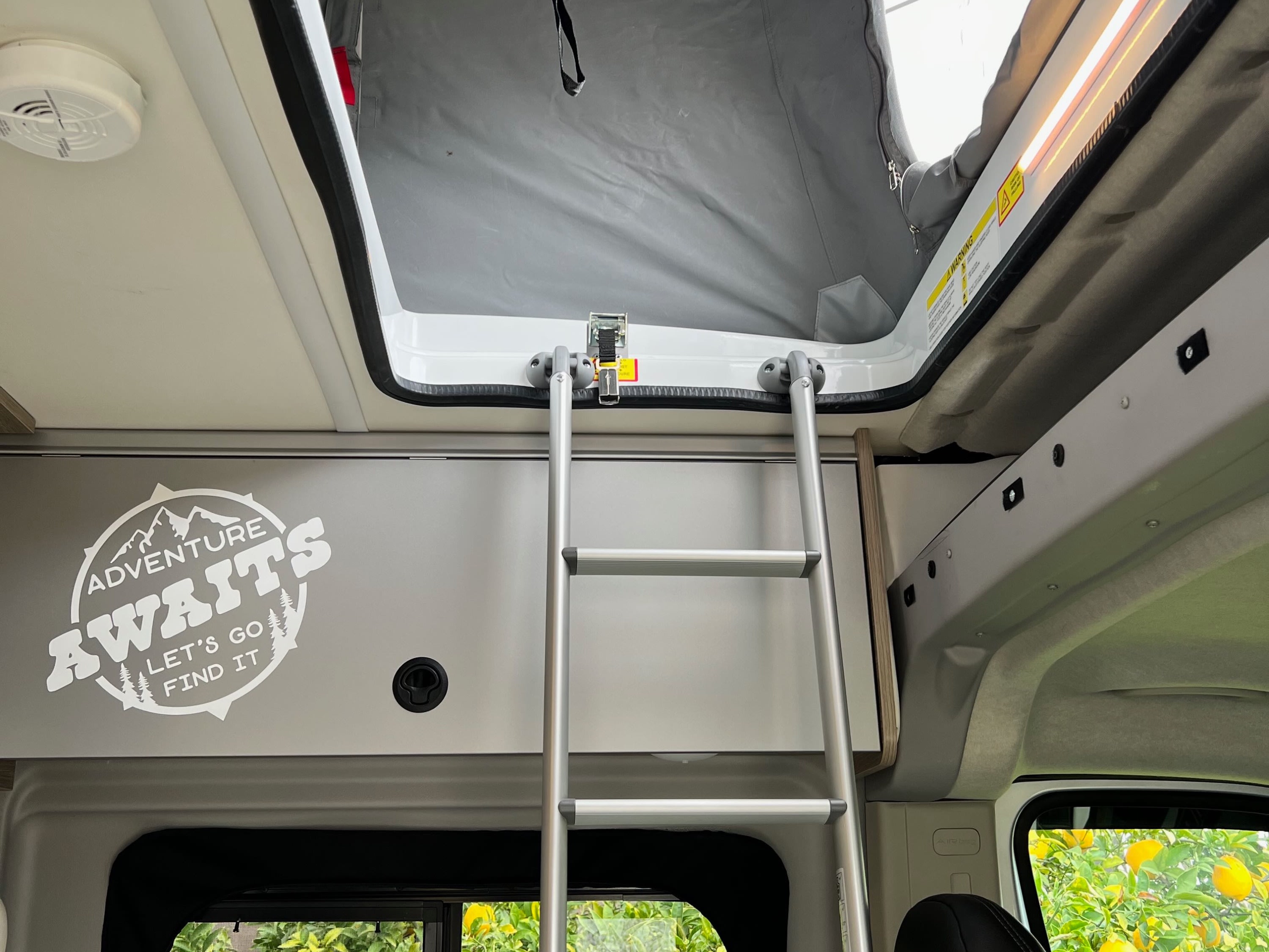 RV rental interior with ladder to pop-top sleeping area. Smoke alarm & 'Adventure Awaits' decal visible. Ready for adventure!. Winnebago Solis 2023