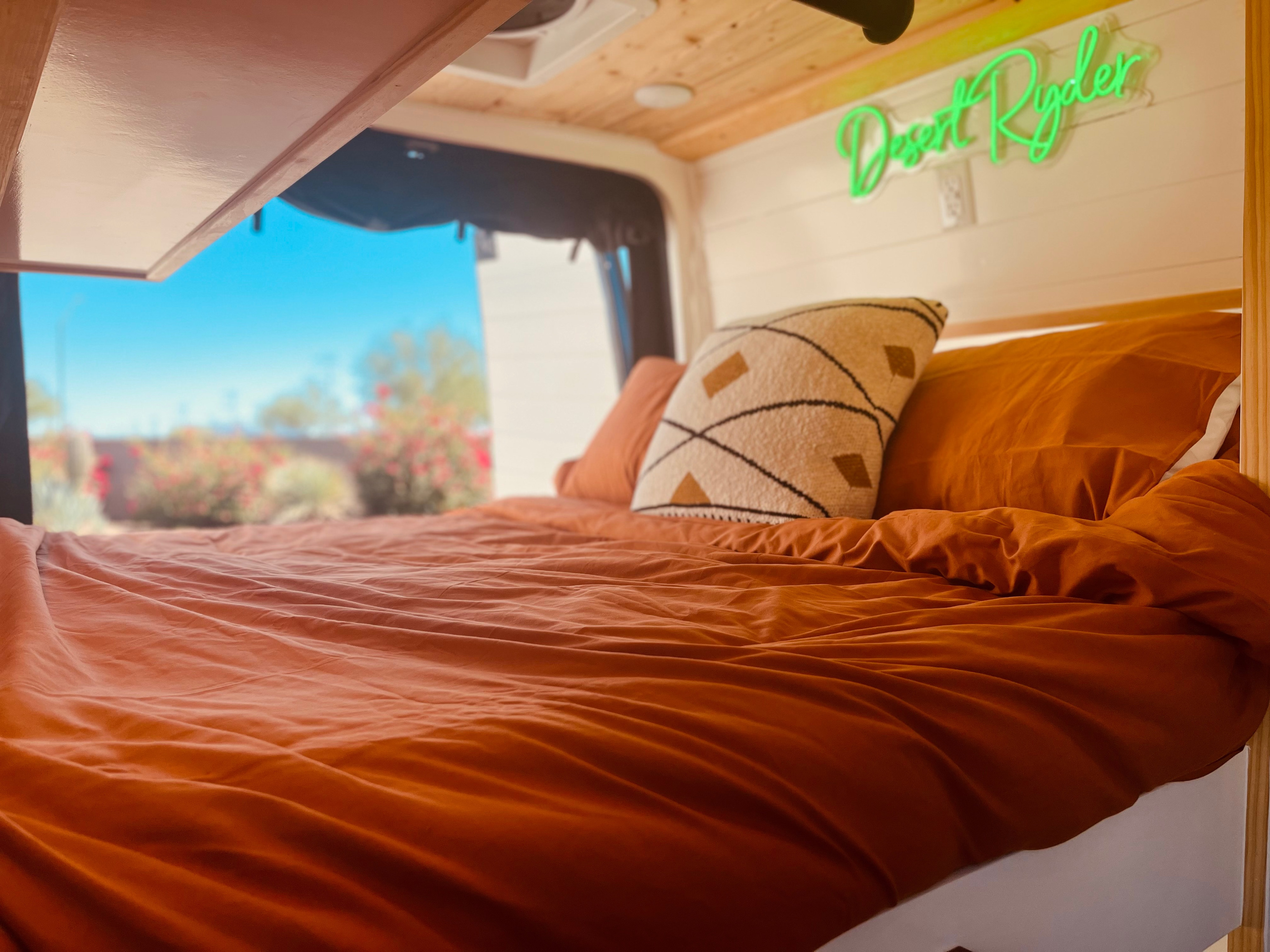 Cozy RV rental van bedroom featuring a burnt orange bed, decorative pillow, "Desert Ryder" neon sign, and a bright outdoor view.. Mercedes Sprinter 2500 2019