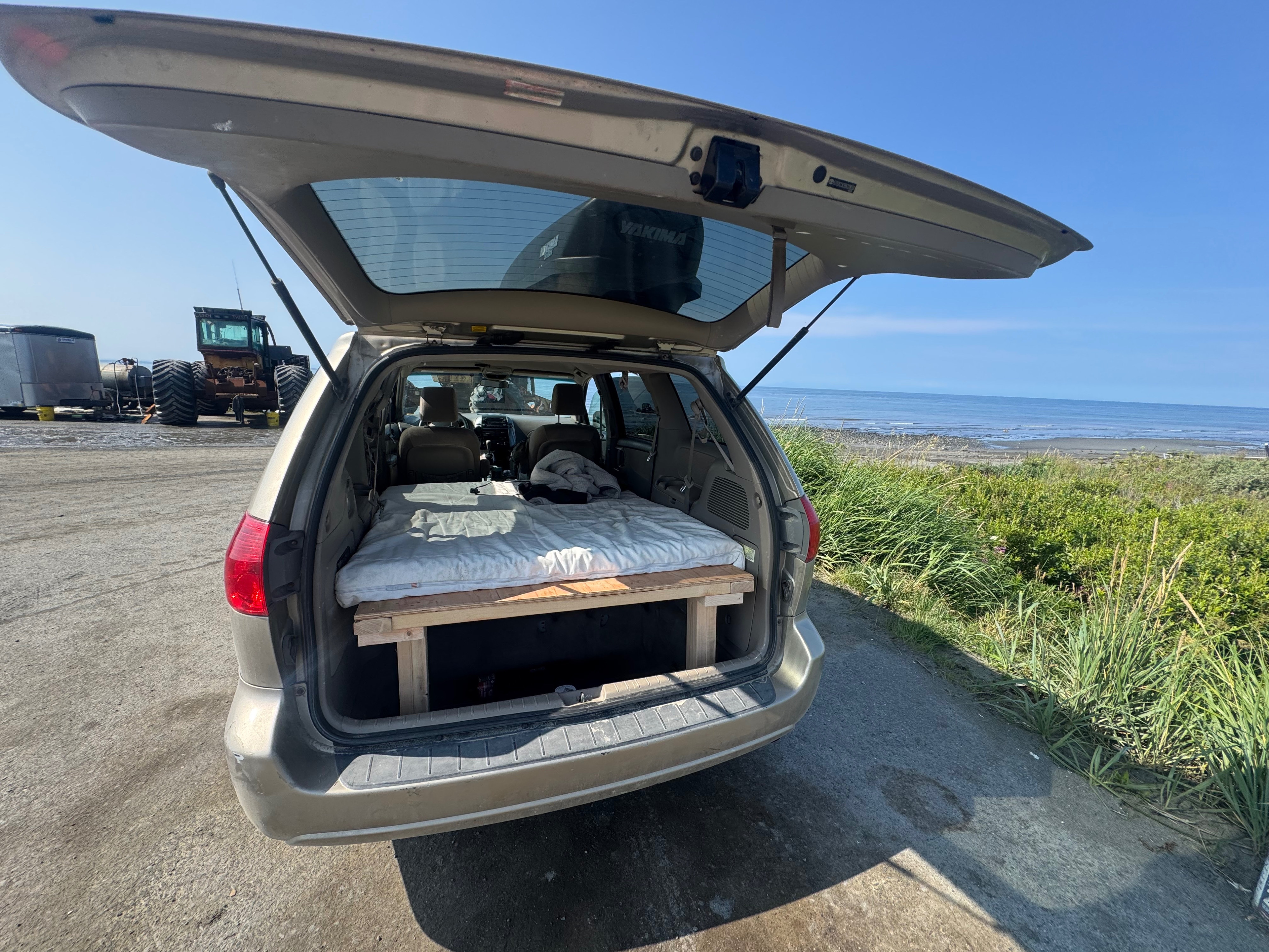 This car, used as an rv rental, has its trunk open, showing a built-in bed by the ocean. A tractor is nearby.. Toyota Sienna 2009