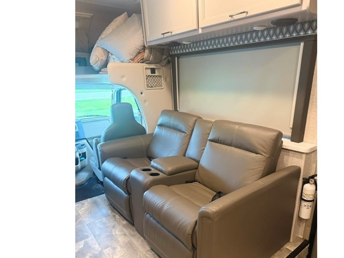 Cozy RV rental interior features dual recliners, an overhead bunk bed, and a window with a shade for comfortable travel.. Thor Quantum 2023