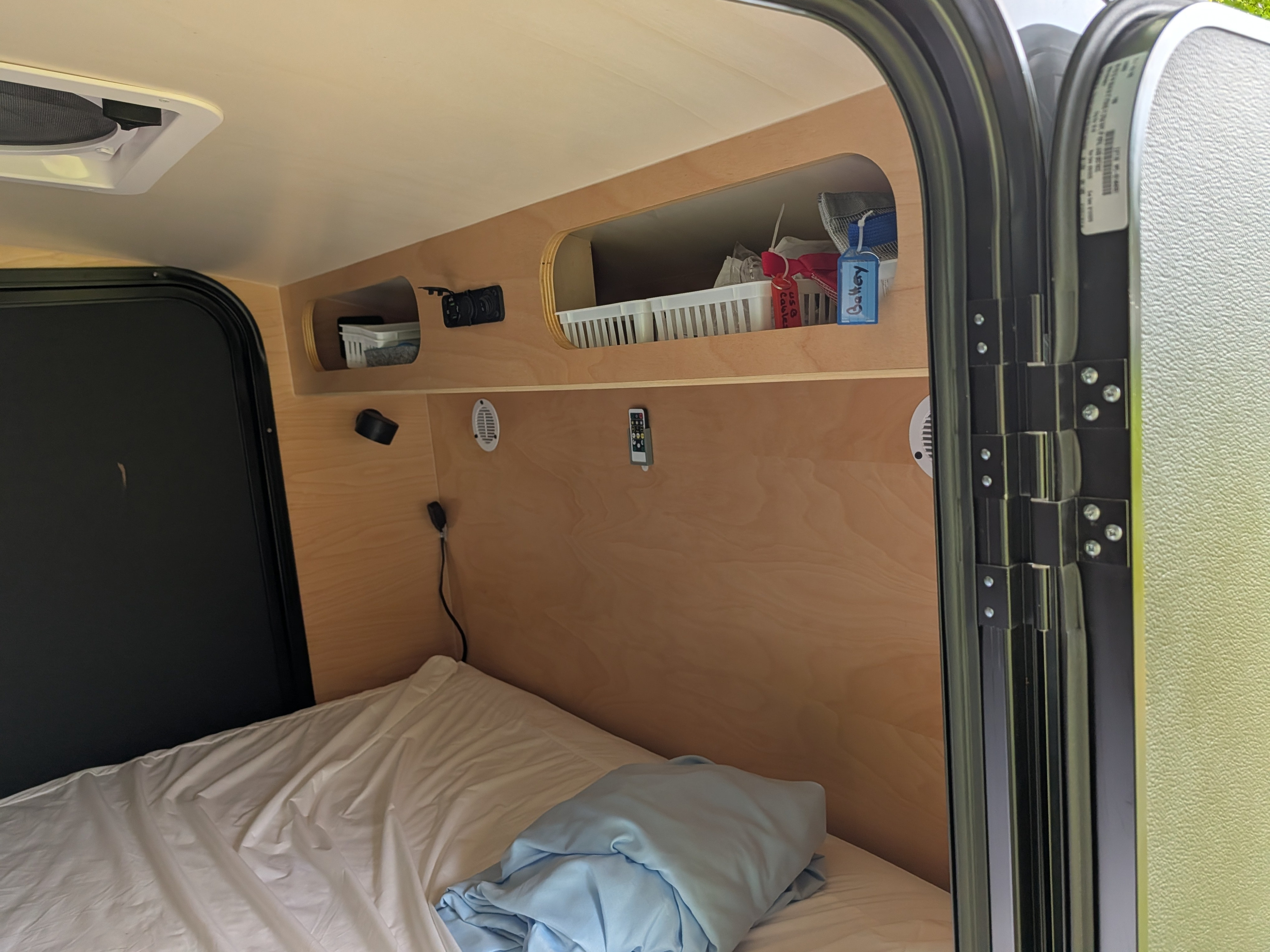 Cozy RV rental interior showing a bed with white sheets & blue blanket, storage cubbies, power outlets, and a ceiling vent.. Hiker Trailer MidRange 5x10 2025