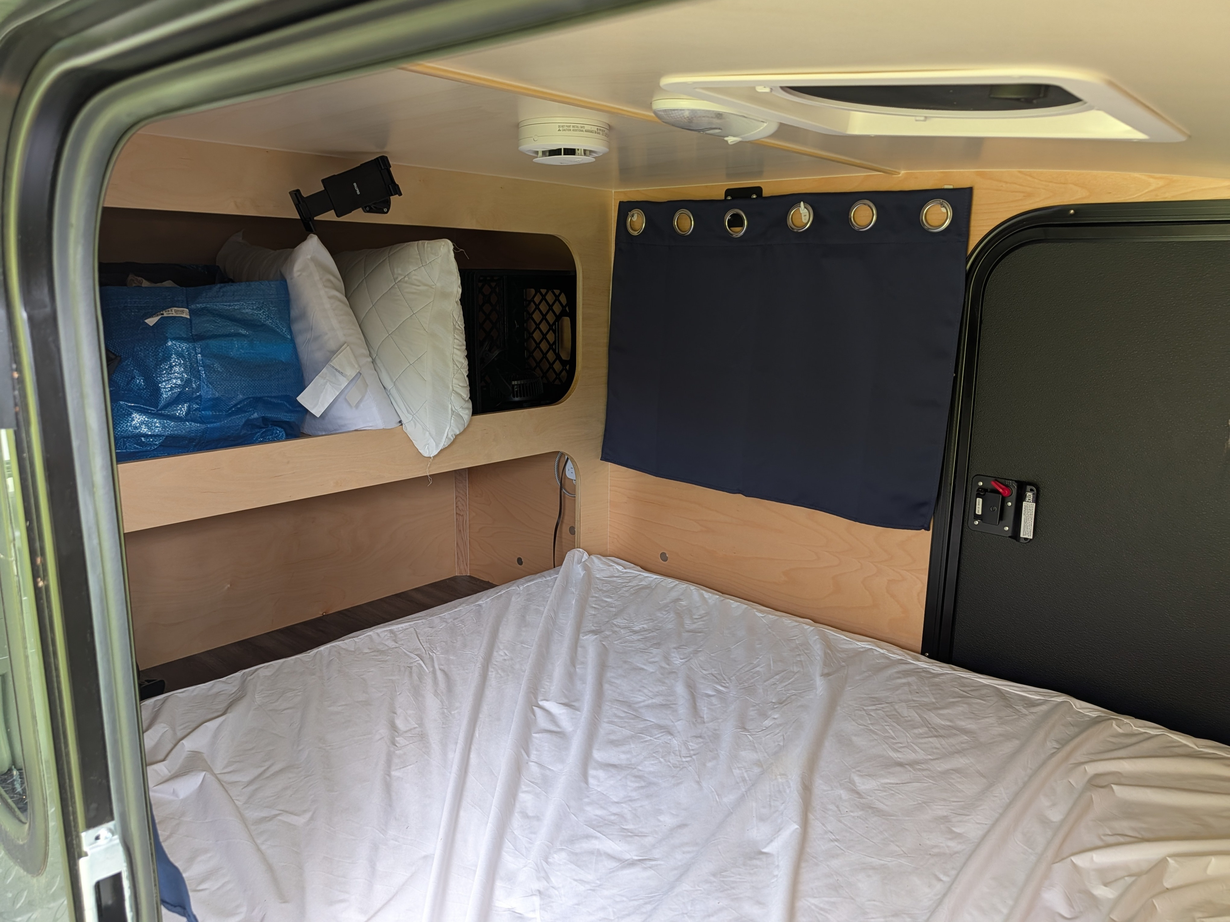 This compact RV rental interior features a bed, overhead storage shelves, a door, and a smoke detector.. Hiker Trailer MidRange 5x10 2025