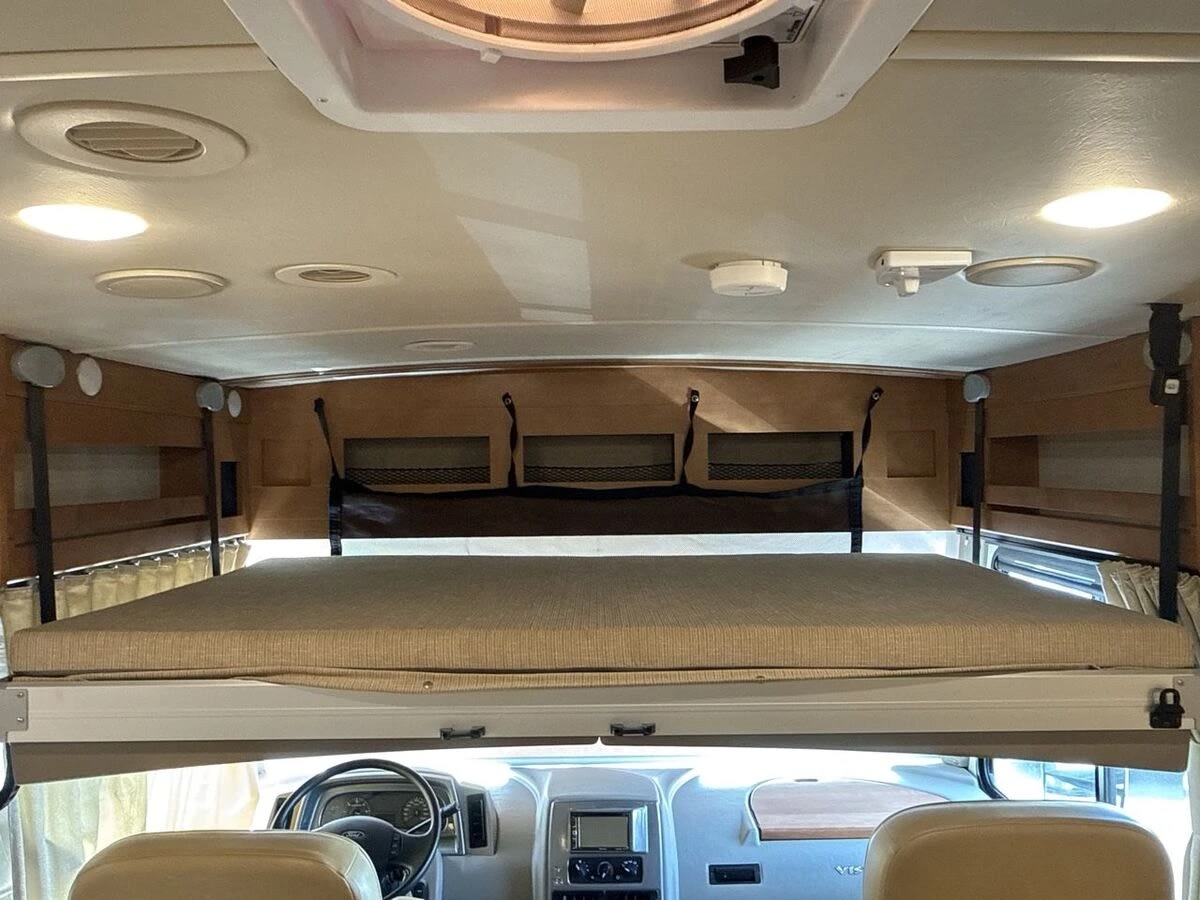 Inside an RV rental, showing the over-cab bunk bed with mattress above the driver's area. Ceiling lights & vents visible.. Winnebago Vista LX 2017