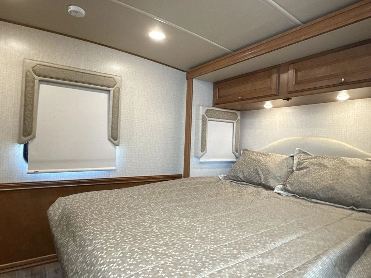 RV rental bedroom showcasing a patterned bed, modern shaded windows, and overhead cabinets with reading lights. Ready for travel!. Winnebago Vista LX 2017