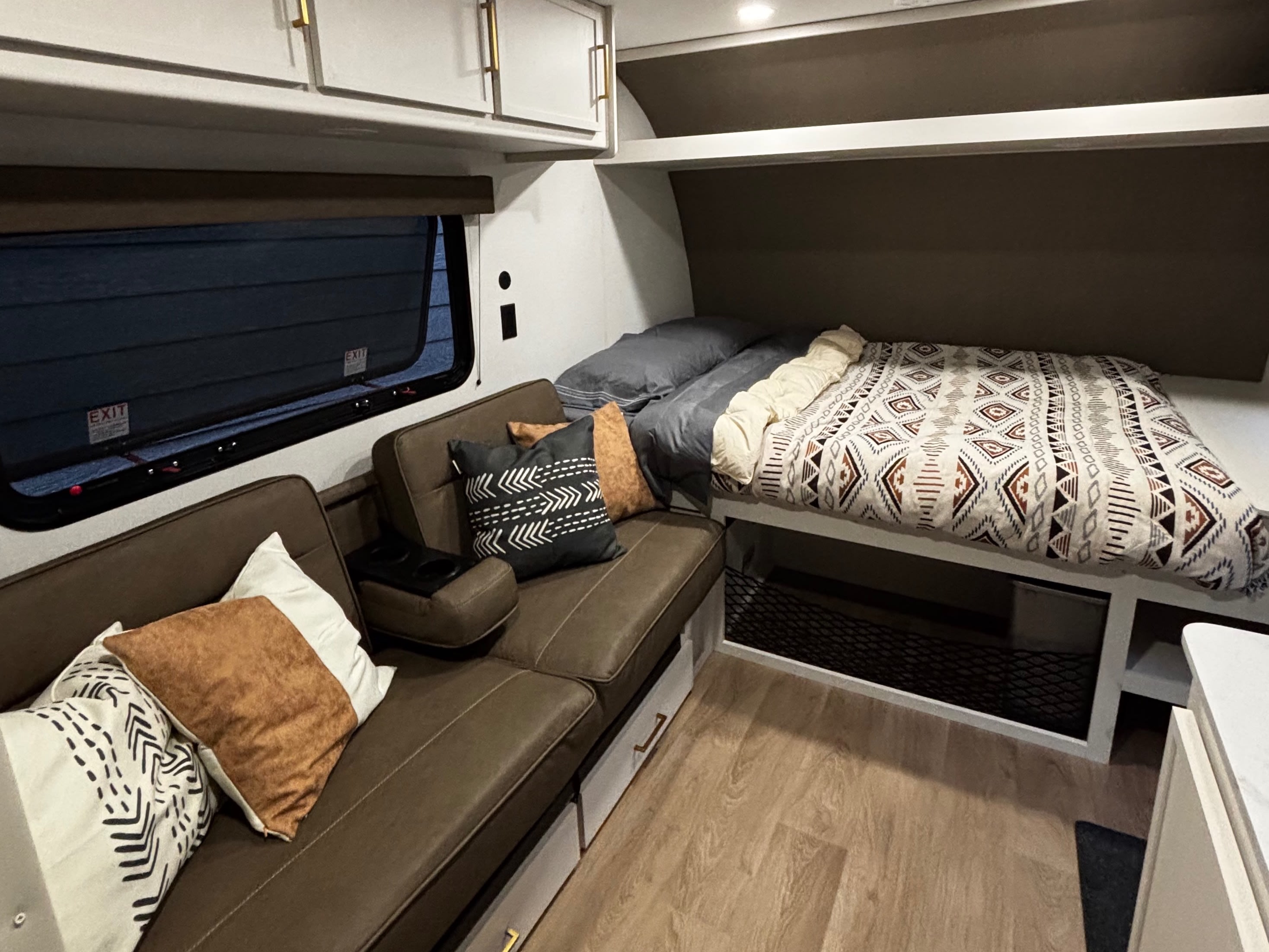 Cozy rv rental interior: brown sofa, bed with patterned blanket, wood-look floor, white cabinets & window.. Grand Design Transcend One 2027