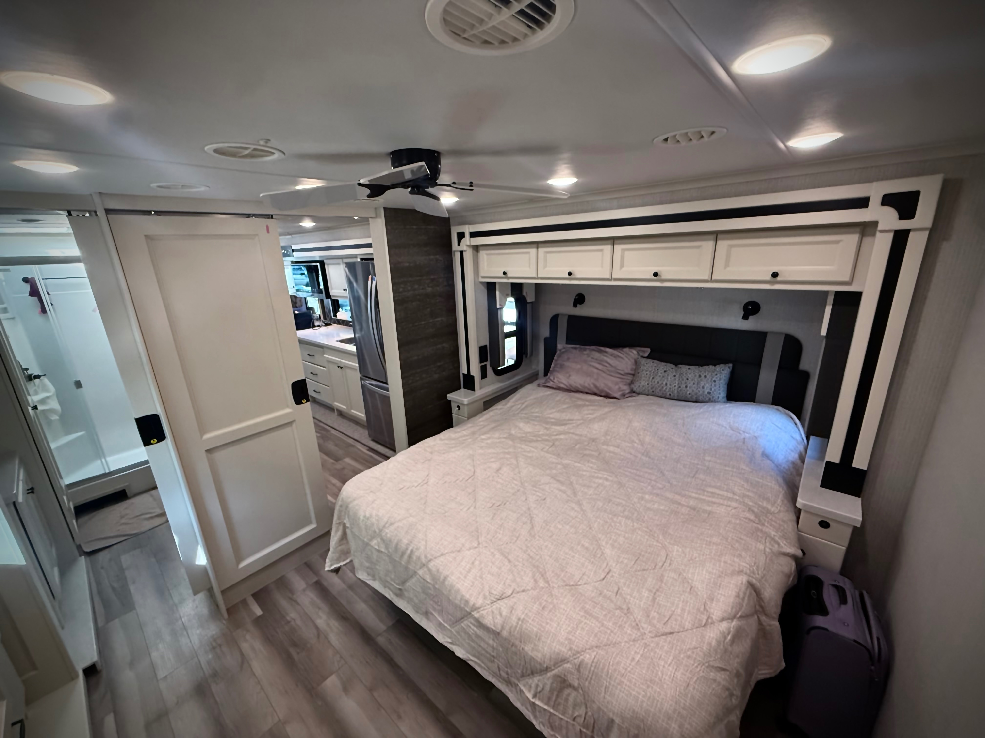 Experience comfort in this RV rental bedroom: large bed, fan, overhead cabinets, and en-suite bath access. Kitchen visible.. Allegro Allegro Open Road 2026