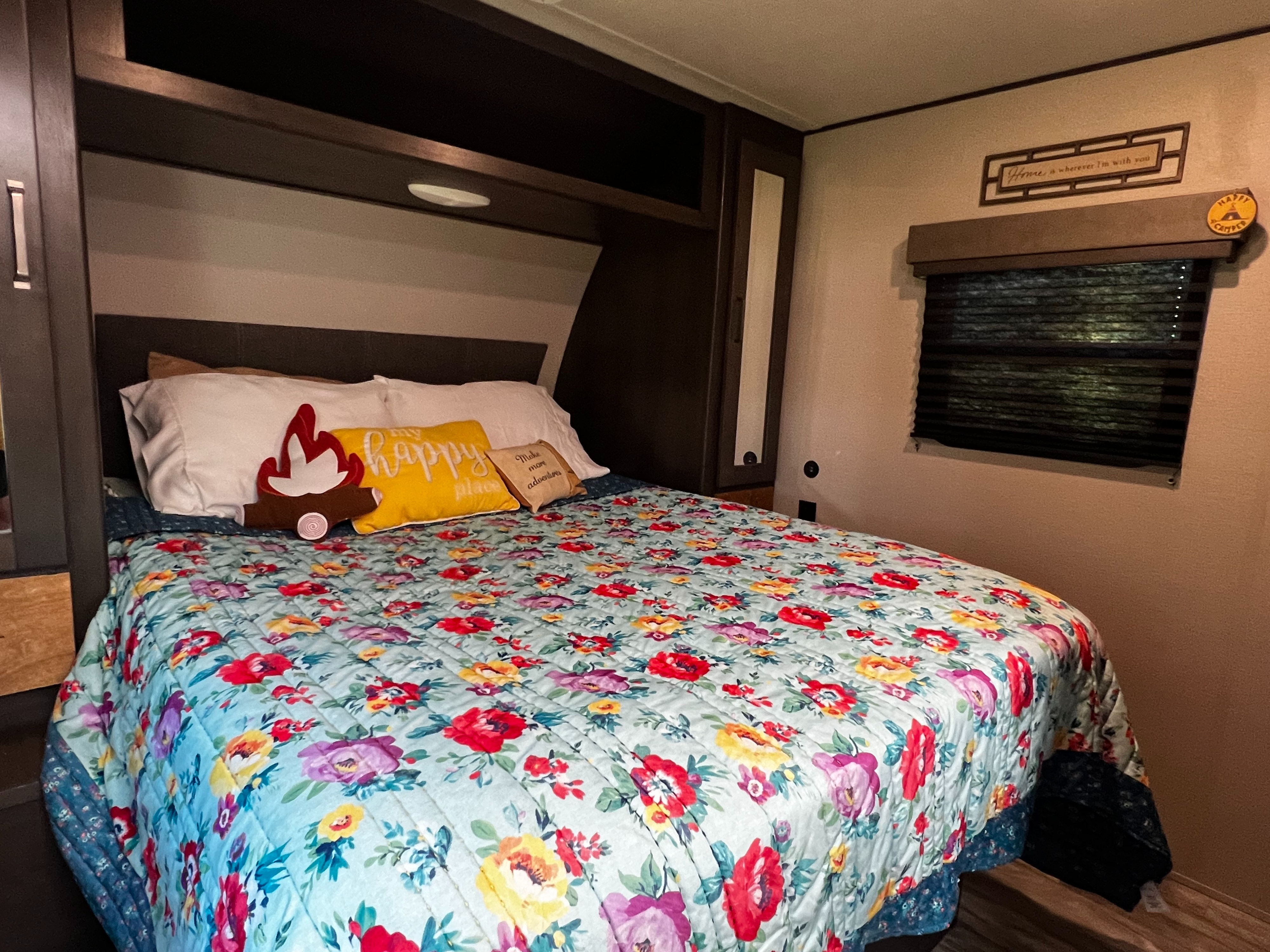 RV rental bedroom: a cozy bed adorned with a vibrant floral quilt, fun themed pillows, and a window with blinds.. Grand Design Transcend Xplor 2021