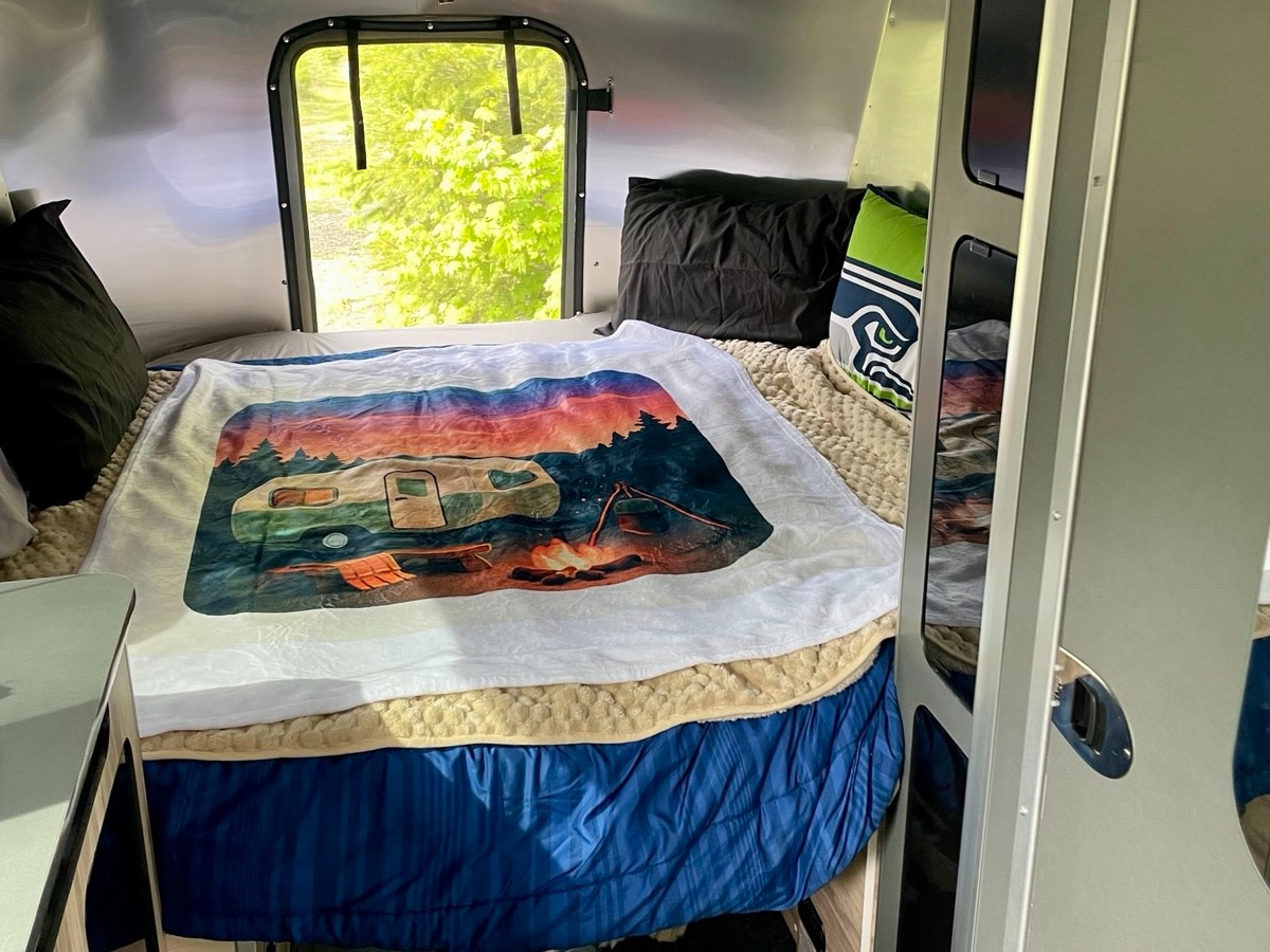 Inside a cozy rv rental: bed with a scenic camper blanket, Seahawks pillow, and window view. Ready for travel!. Airstream Basecamp 2023