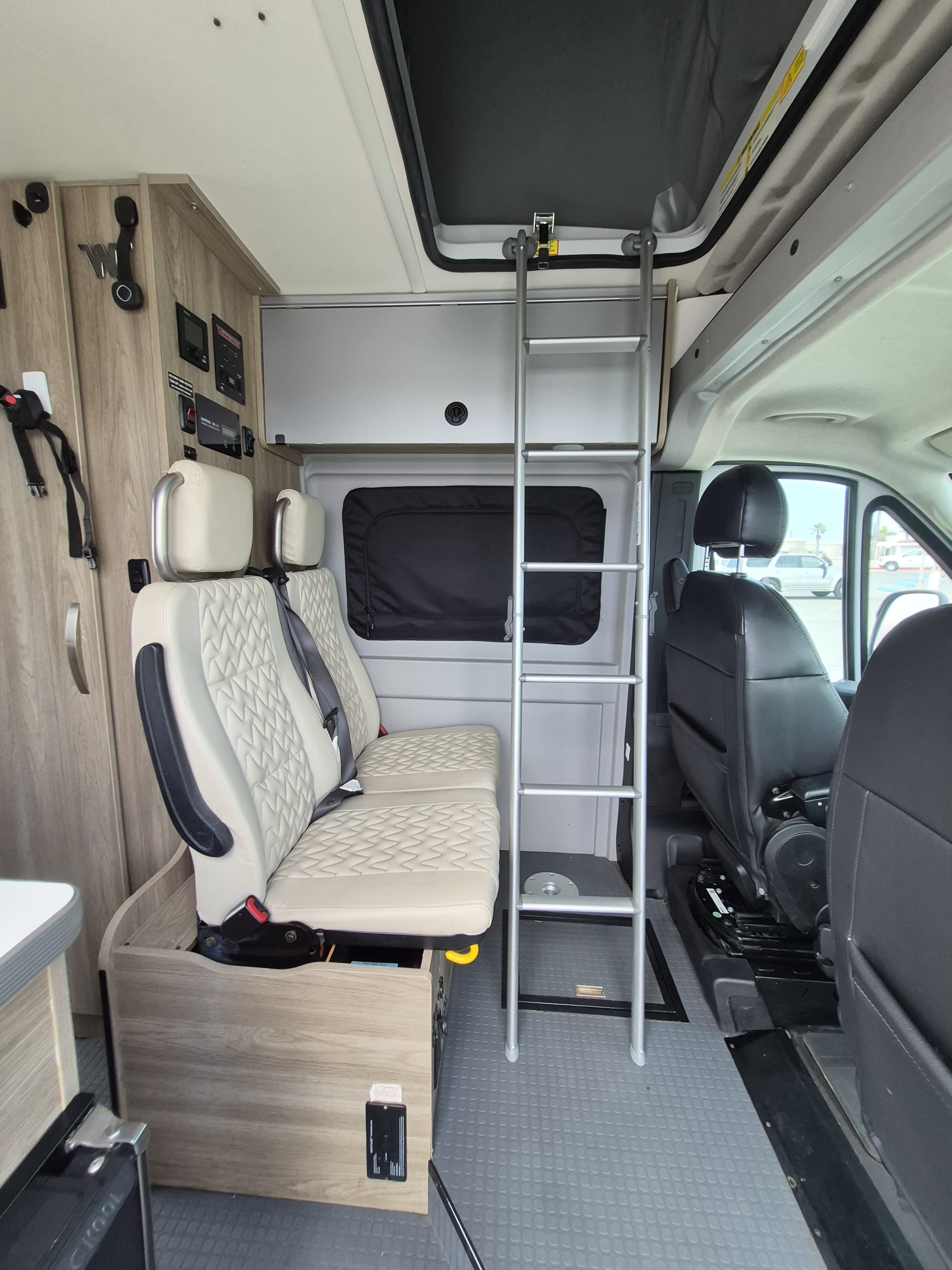 Inside an RV rental, featuring cream passenger seats, a control panel, and a ladder leading to the upper sleeping area.. Winnebago Solis 2023