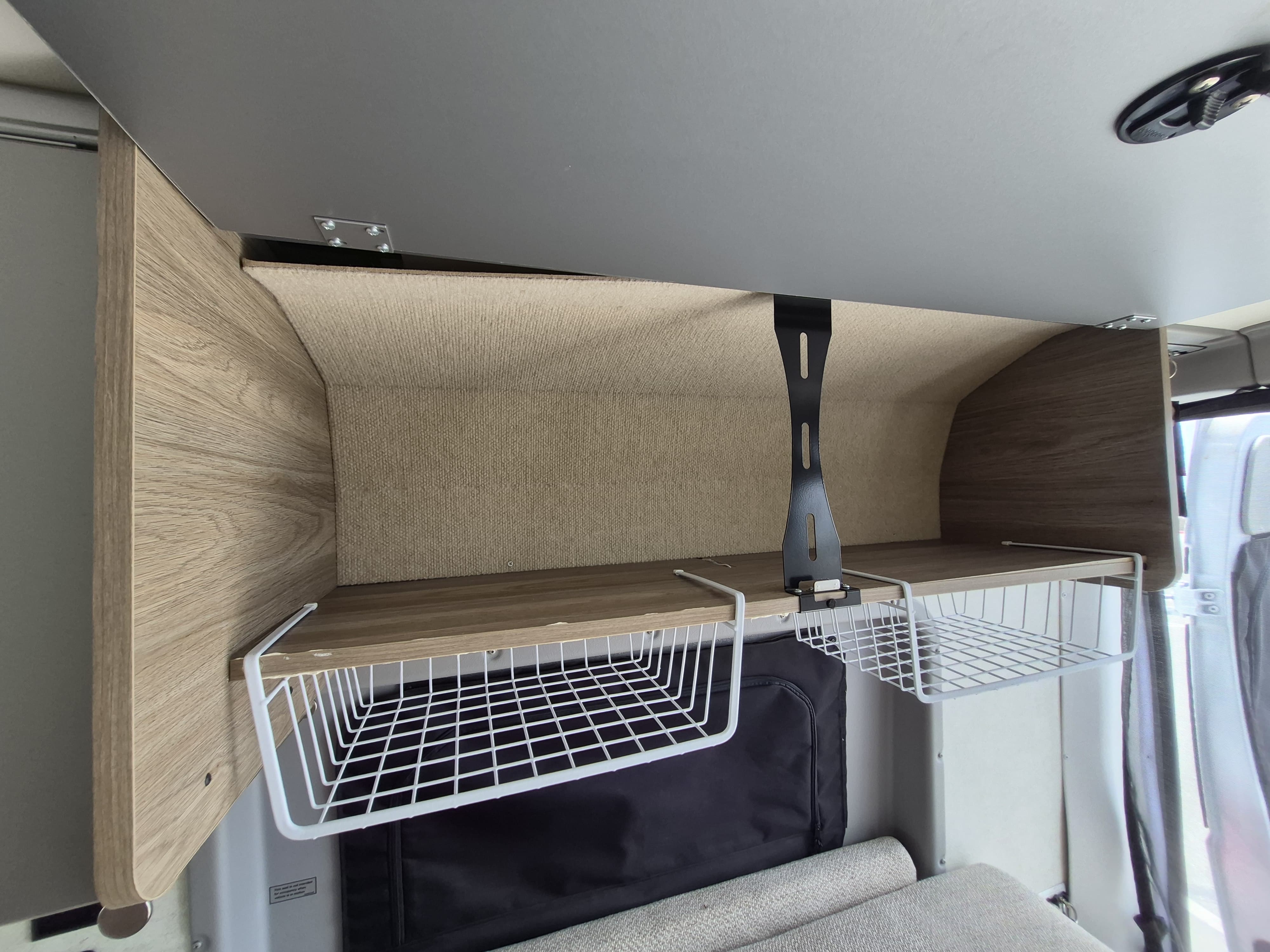 RV rental interior: Overhead wooden shelf with beige fabric lining and two white hanging wire baskets for storage.. Winnebago Solis 2023