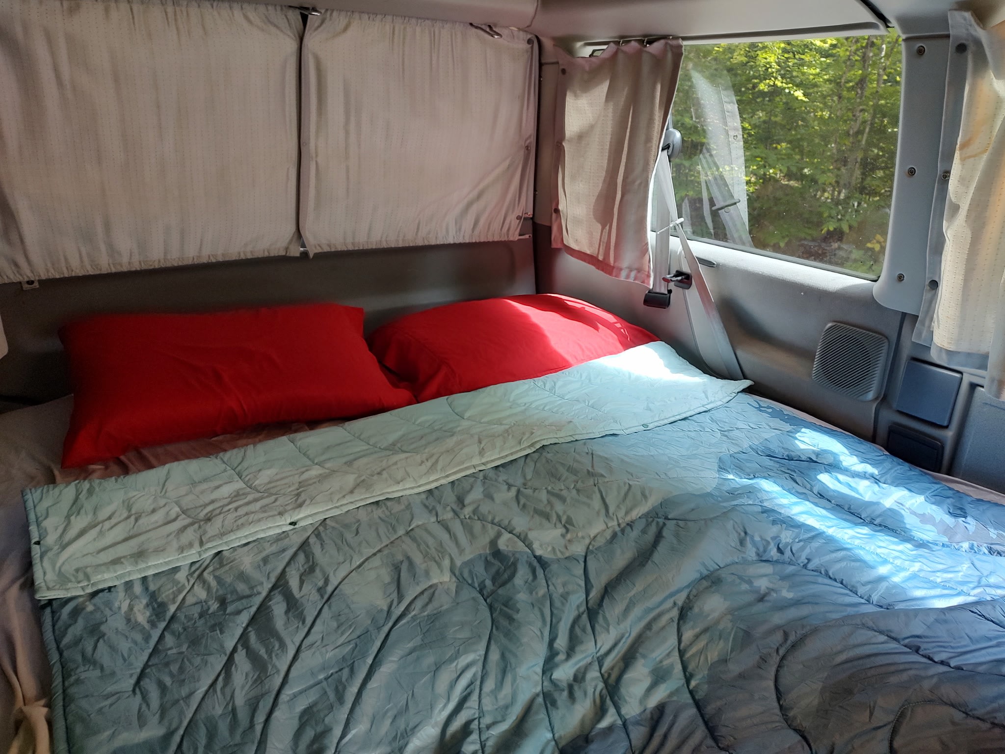Cozy sleeping area in an rv rental van, featuring red pillows, blue comforter, and windows overlooking nature. Ideal for road trips.. Volkswagen T3 Westfalia 2001
