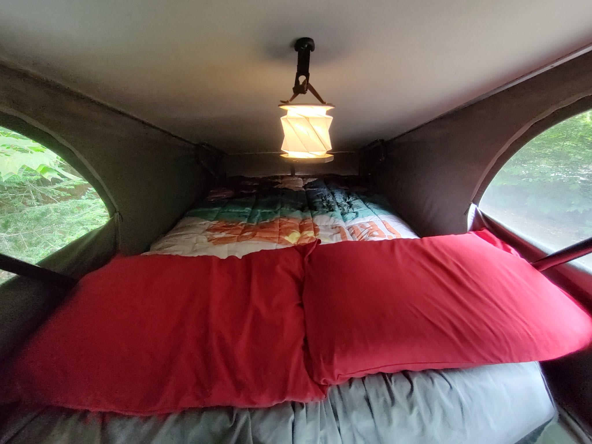 RV rental interior: cozy sleeping area with red pillows, colorful quilt, and hanging light. Nature visible outside.. Volkswagen T3 Westfalia 2001
