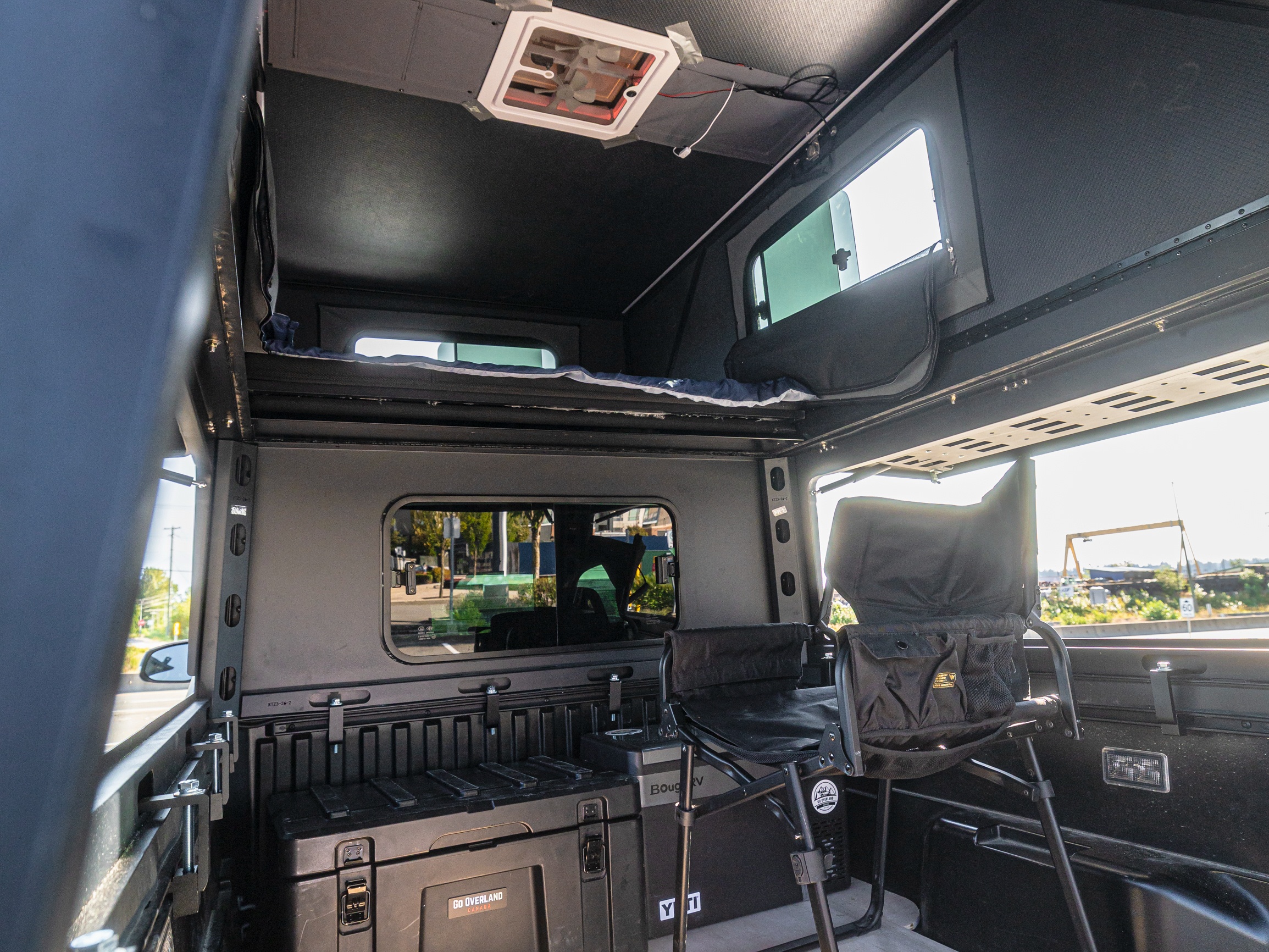 Inside a modular truck camper, an rv rental adventure setup. Features elevated bed, ceiling fan, storage boxes, and camp chair.. Toyota Tacoma 2025