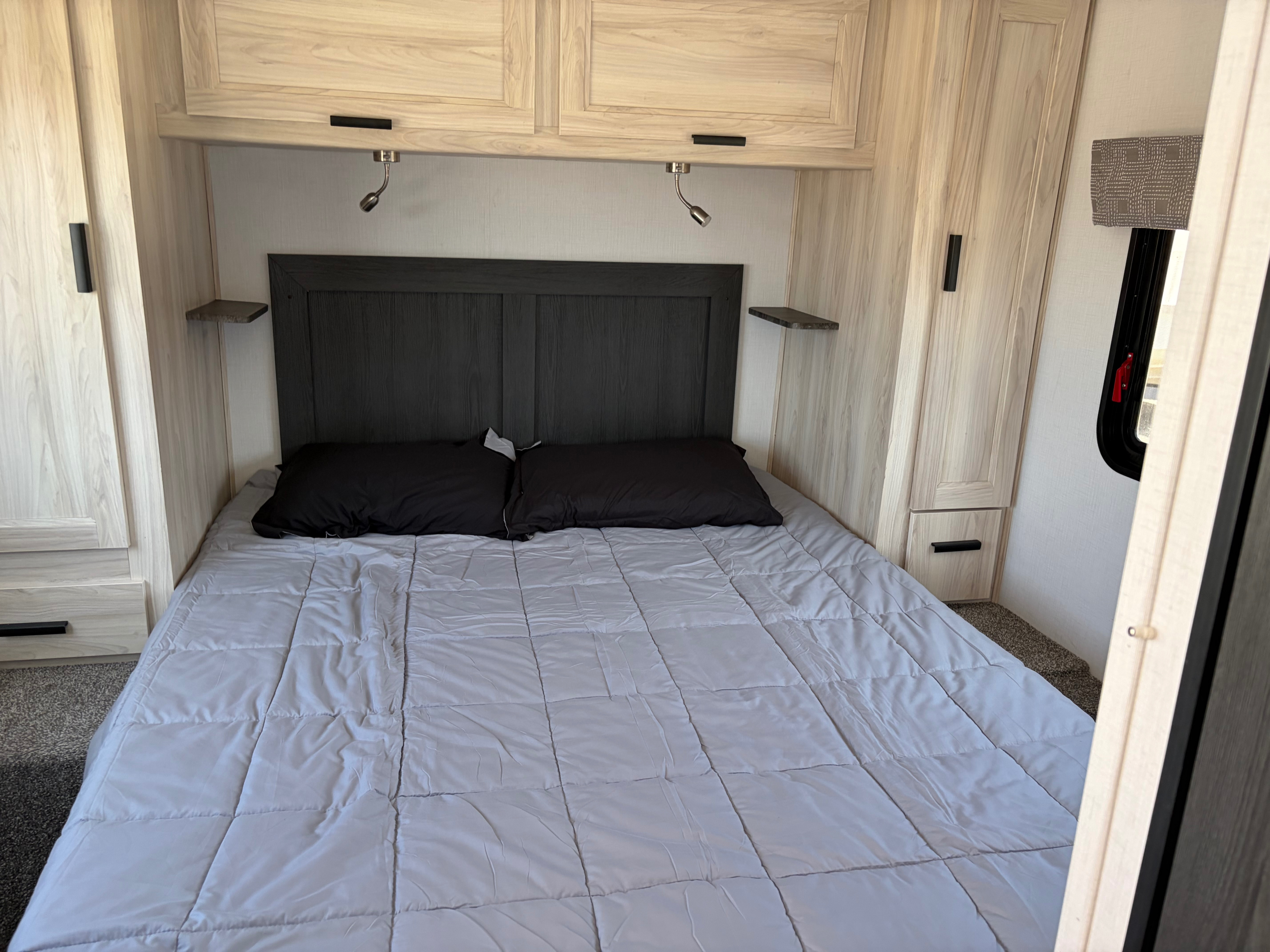 Cozy RV rental bedroom featuring a full bed with a gray comforter, dark headboard, light wood cabinetry, reading lights, and side storage.. Forest River Sunseeker LE 2024