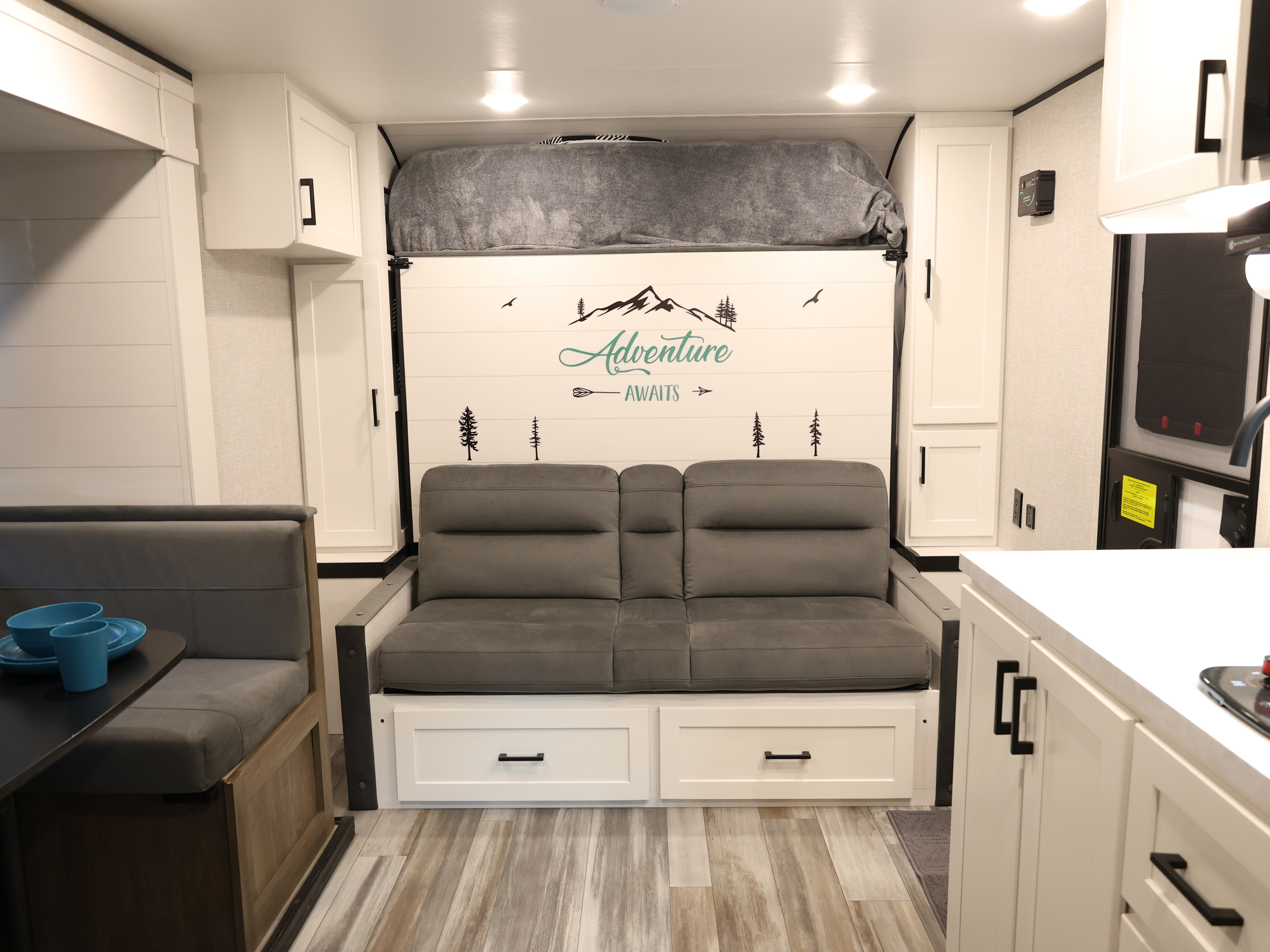 Functional rv rental interior with a fold-down bed above a sofa, a dinette, and white cabinetry. "Adventure Awaits" wall decal.. Jayco Jay Flight SLX Baja Edition 2025