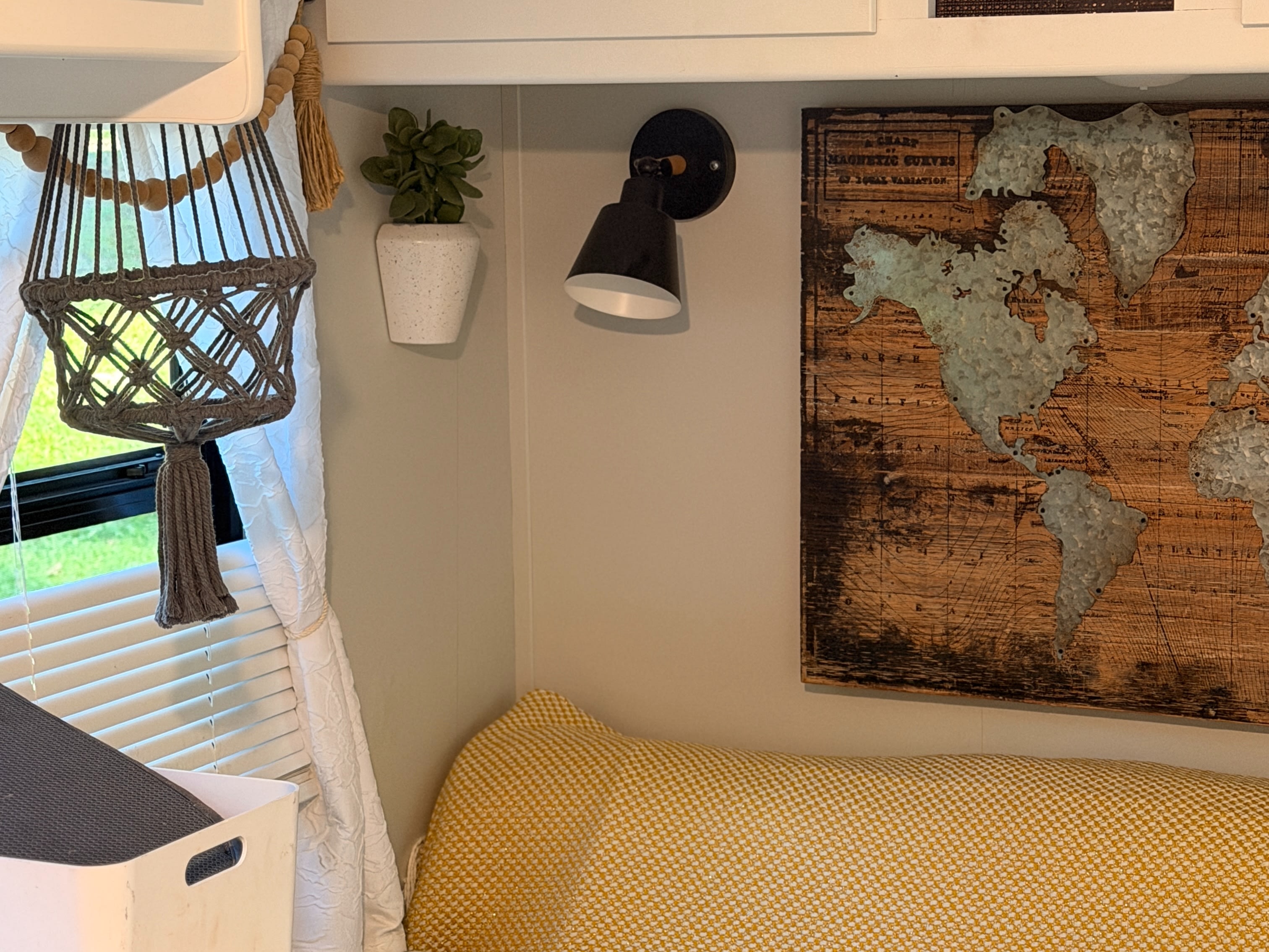 Cozy RV rental interior: macrame hanger, wall plant, reading light, and a world map above a yellow cushion.. Clipper Cadet 2021