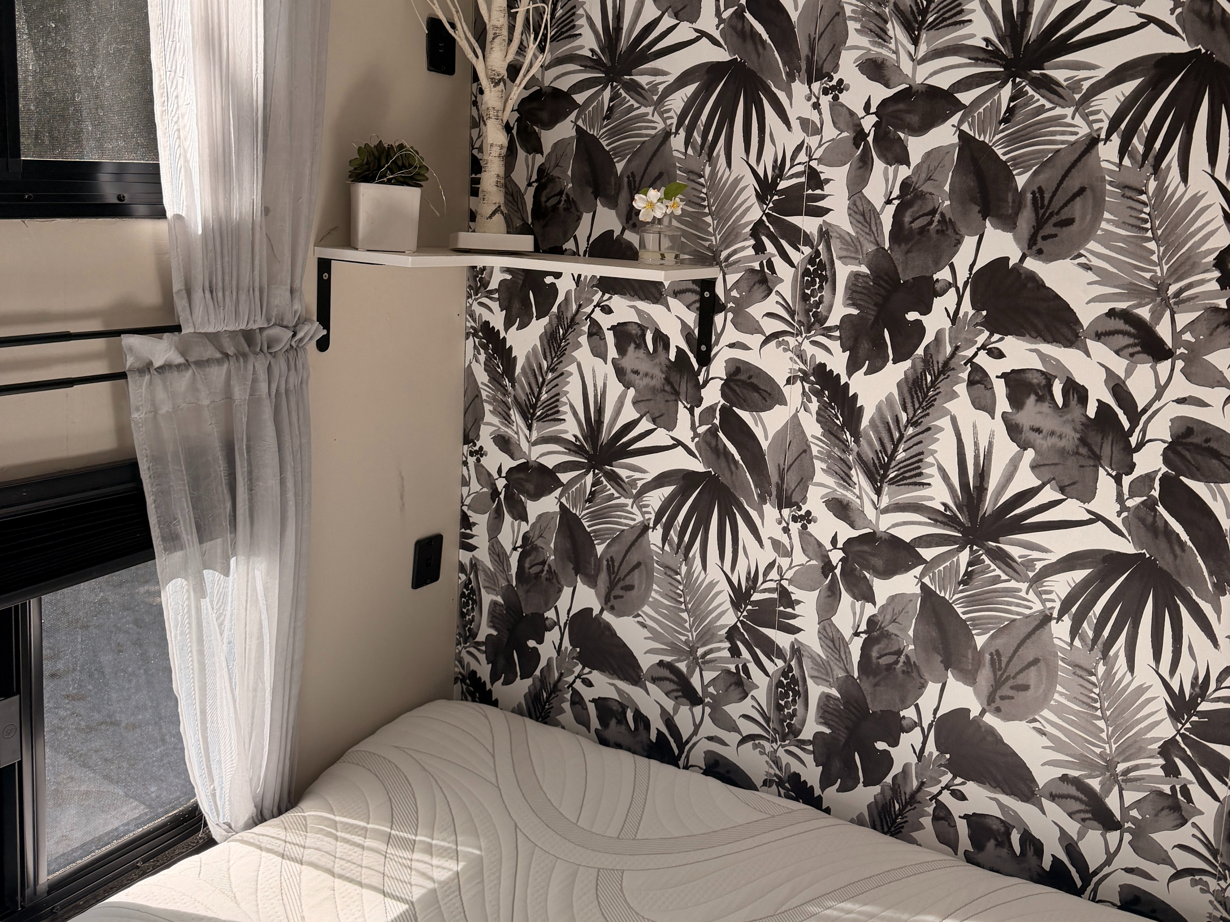 Compact RV rental bedroom featuring a bed, black & white tropical wallpaper, a shelf with decor, and a sheer curtained window.. Clipper Cadet 2021