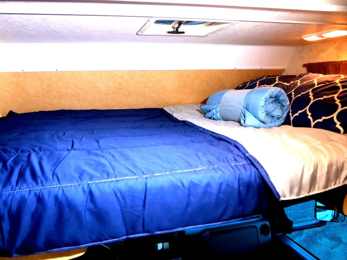Cozy RV rental bed setup with a blue comforter, patterned pillows, and a rolled-up blanket in the overhead bunk.. Thor Majestic 2016