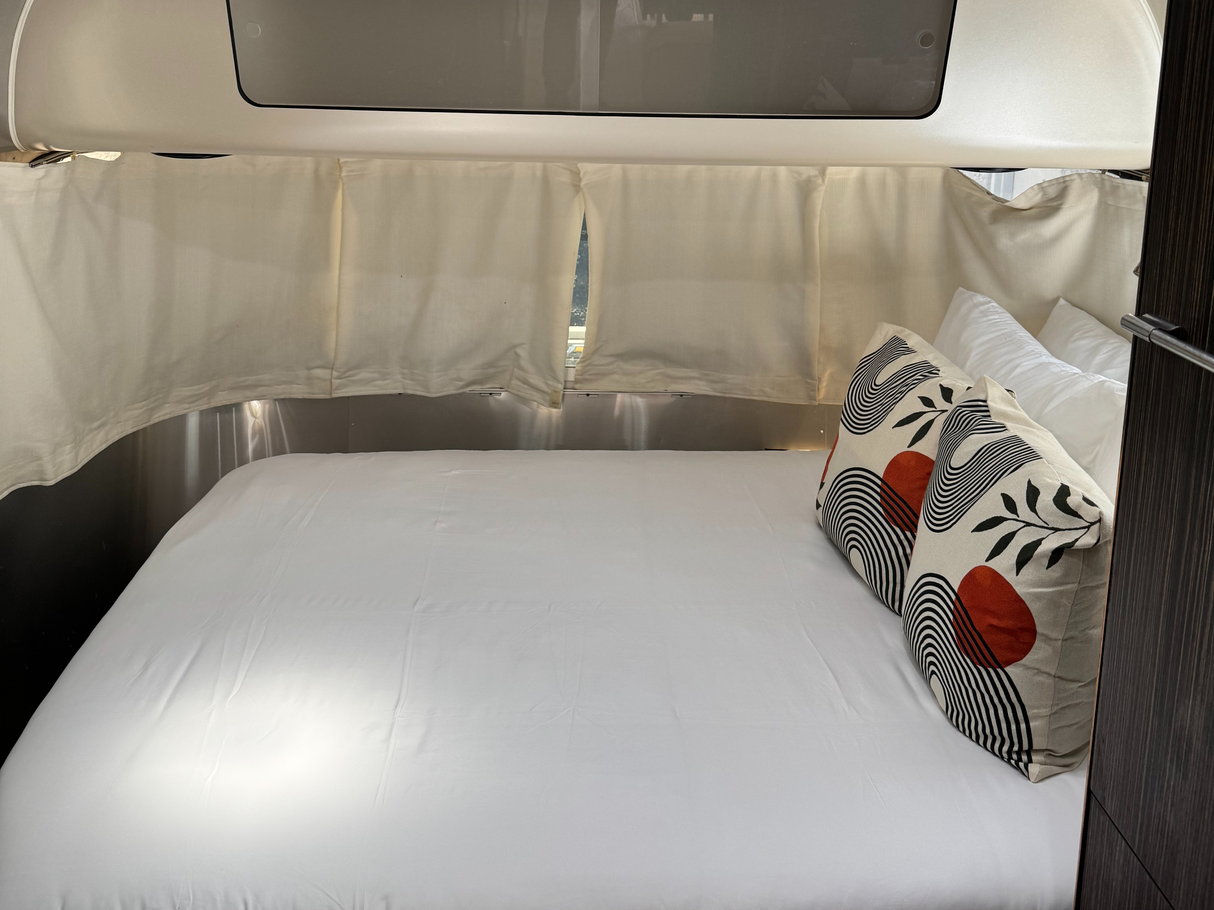 A cozy RV rental bedroom interior, featuring a clean white bed with modern patterned pillows and privacy curtains.. Airstream International 2017