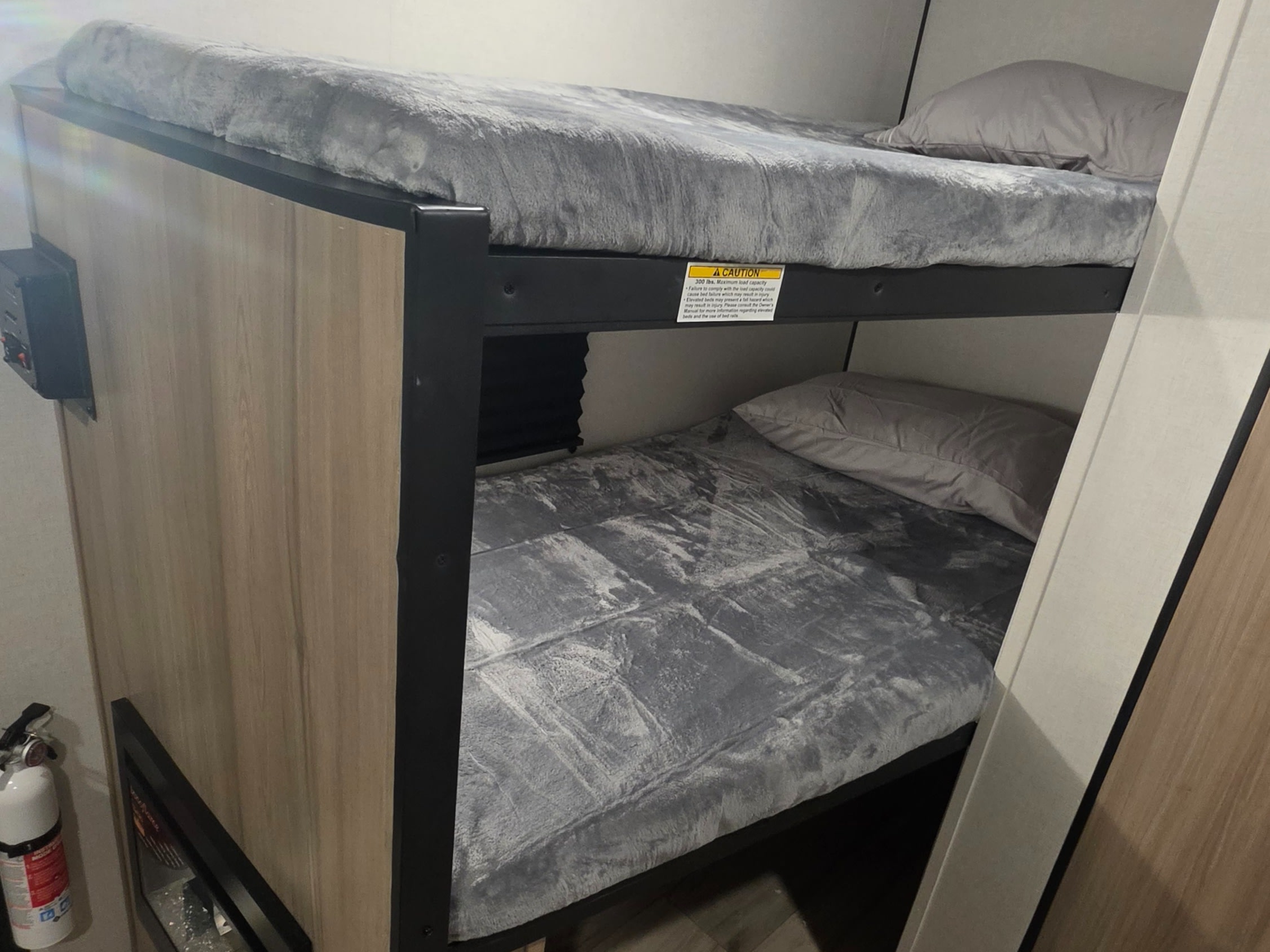 RV rental bunk beds feature cozy grey bedding. A safety sticker warns of 300 lbs max capacity. A fire extinguisher is nearby.. Keystone RV Coleman 2026