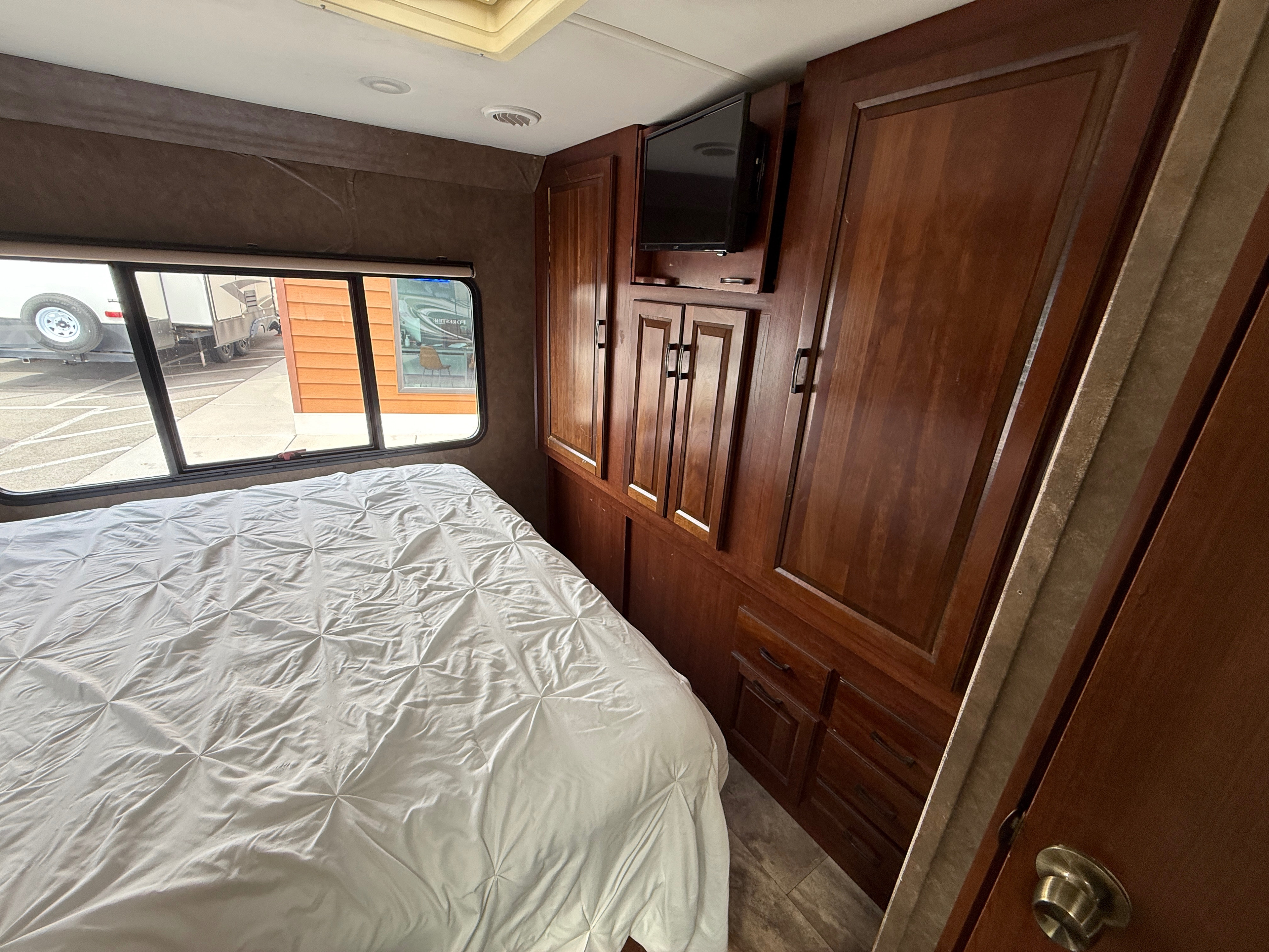 Cozy RV rental bedroom with a white bed, storage cabinets, TV, and a window looking out at other RVs.. Forester Forester Motorhome 2016