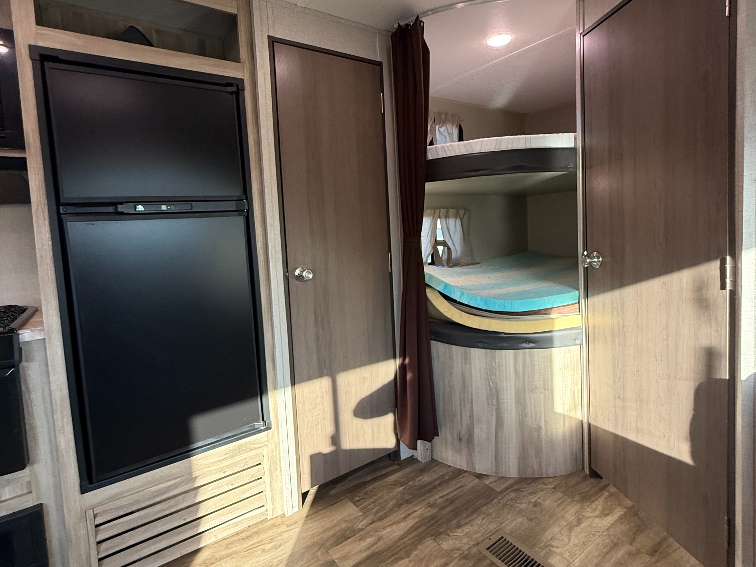 RV rental interior: compact refrigerator, a wooden door, and cozy bunk beds with colorful foam mattresses in a modern camper.. Coachmen Catalina Summit Series 261BHS 2020