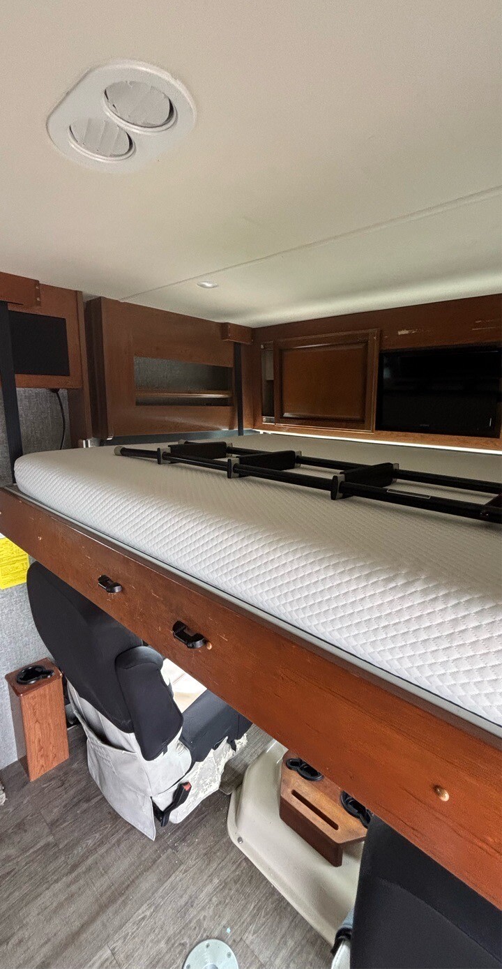 RV rental interior showing a drop-down bunk bed with white mattress & safety netting, overhead AC, and wood trim.. Fleetwood Bounder 2018