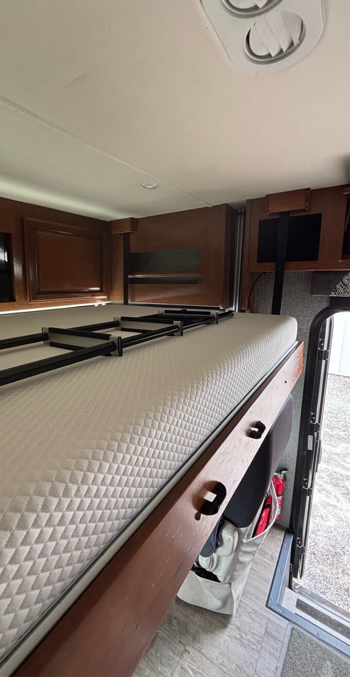 Inside an RV rental: a bunk bed with a white quilted mattress and a black folded ladder, surrounded by wood trim. An open door shows outside.. Fleetwood Bounder 2018