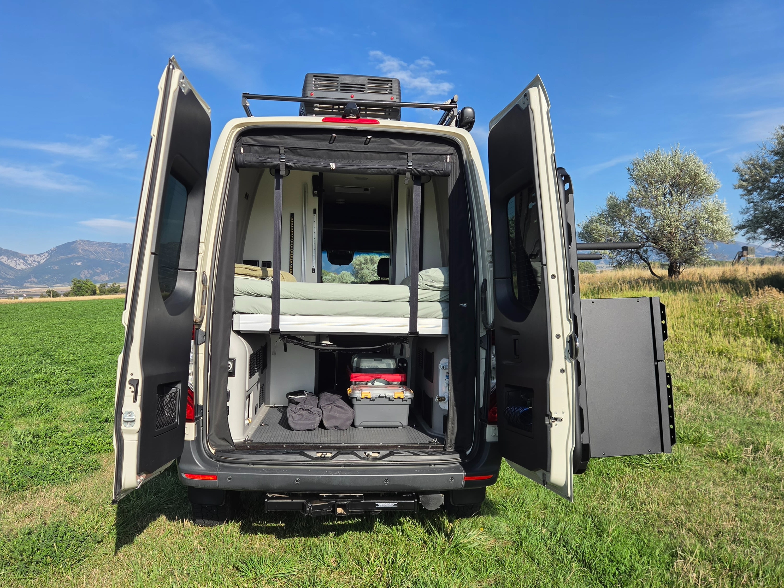 This rv rental conversion van sits in a field, rear doors open to reveal an elevated bed, storage, and mountain views.. Winnebago Revel 2024