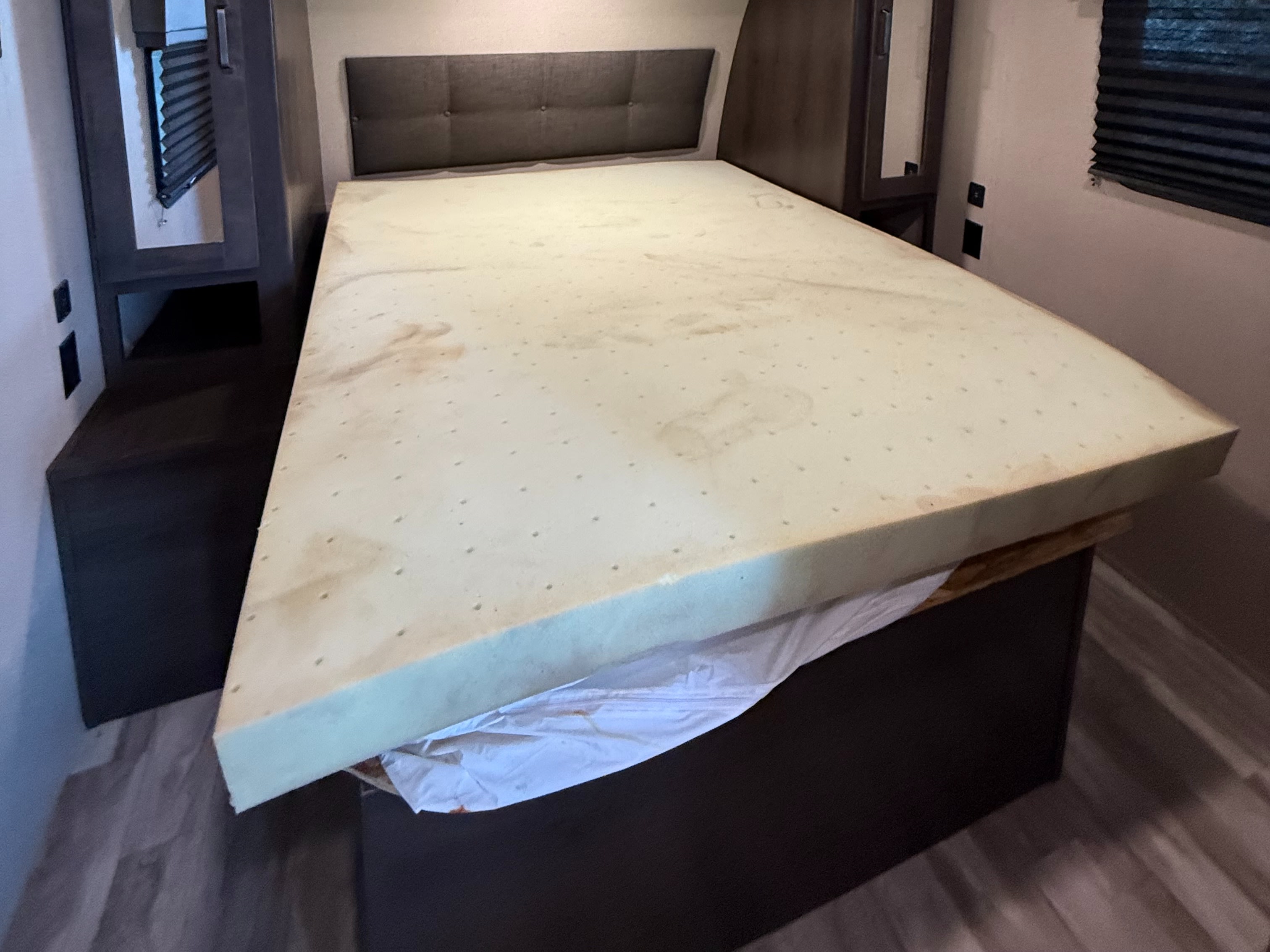 This RV rental bedroom features a worn, stained, and perforated foam mattress on a bed with a dark headboard and built-in storage.. Grand Design Transcend Xplor 2022
