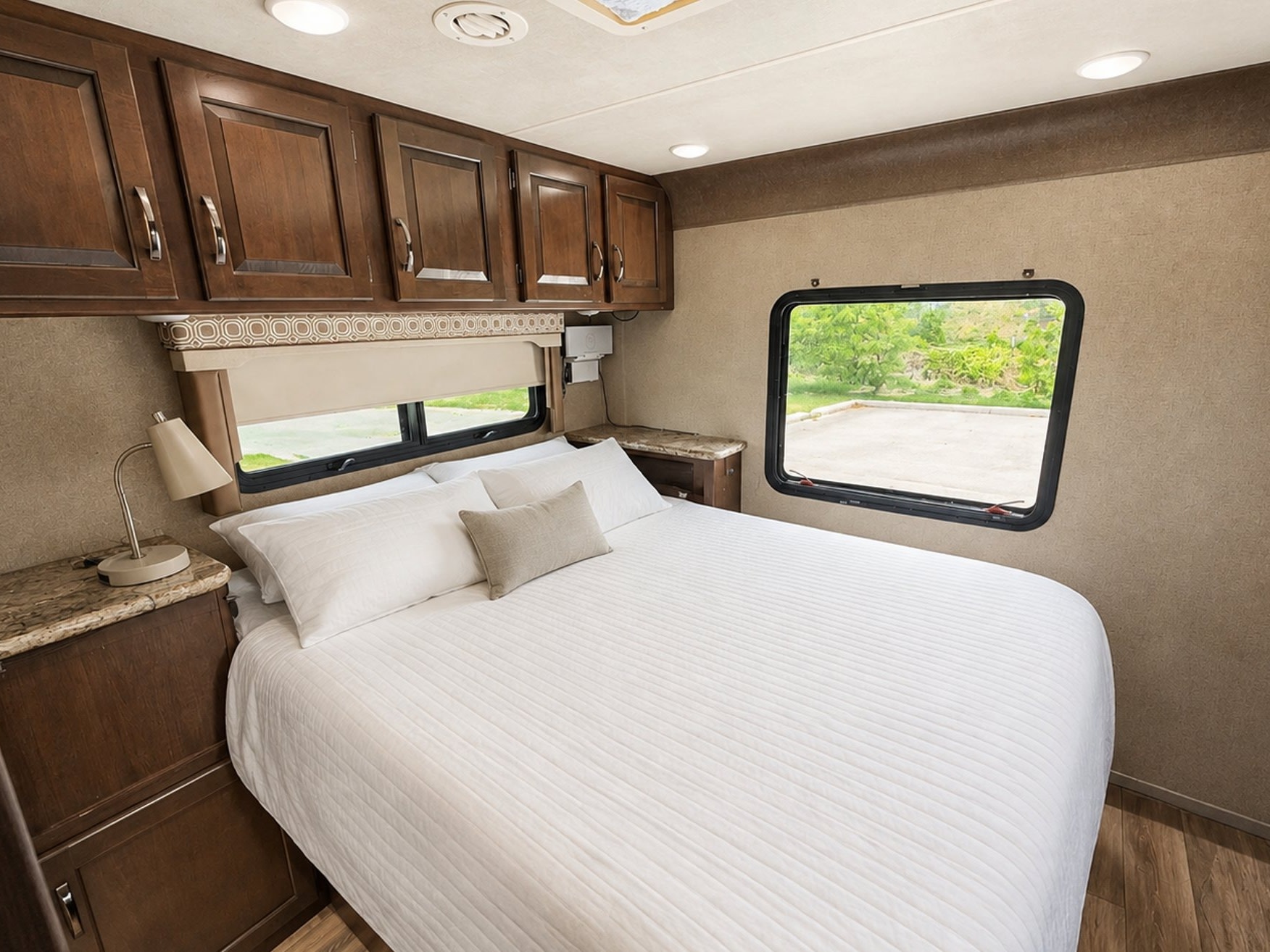 Cozy RV rental bedroom featuring a comfortable queen bed with white bedding, wooden overhead cabinets, and windows with outdoor views.. Thor Four Winds 2020