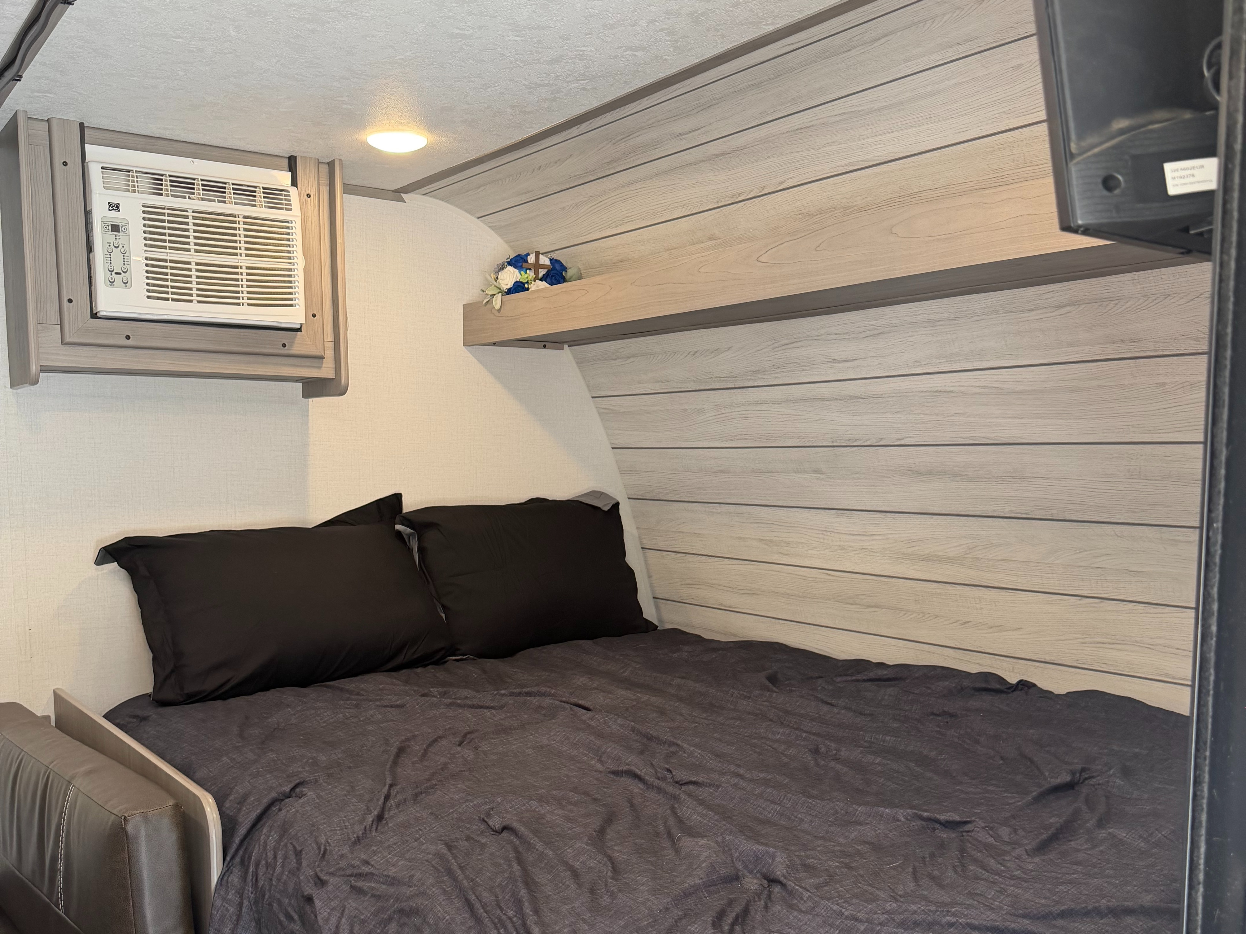 Inside an RV rental: a comfy bed with dark linens, wall-mounted AC, and a decorative shelf. Ideal for rest on the road.. Keystone RV Coleman Lantern LT 17B 2024