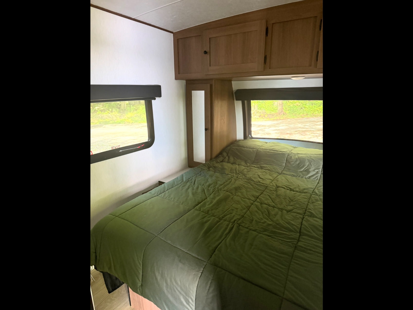 Cozy RV rental bedroom featuring a bed with a green comforter, windows revealing nature, and overhead storage cabinets.. Coachmen Apex Nano 2026