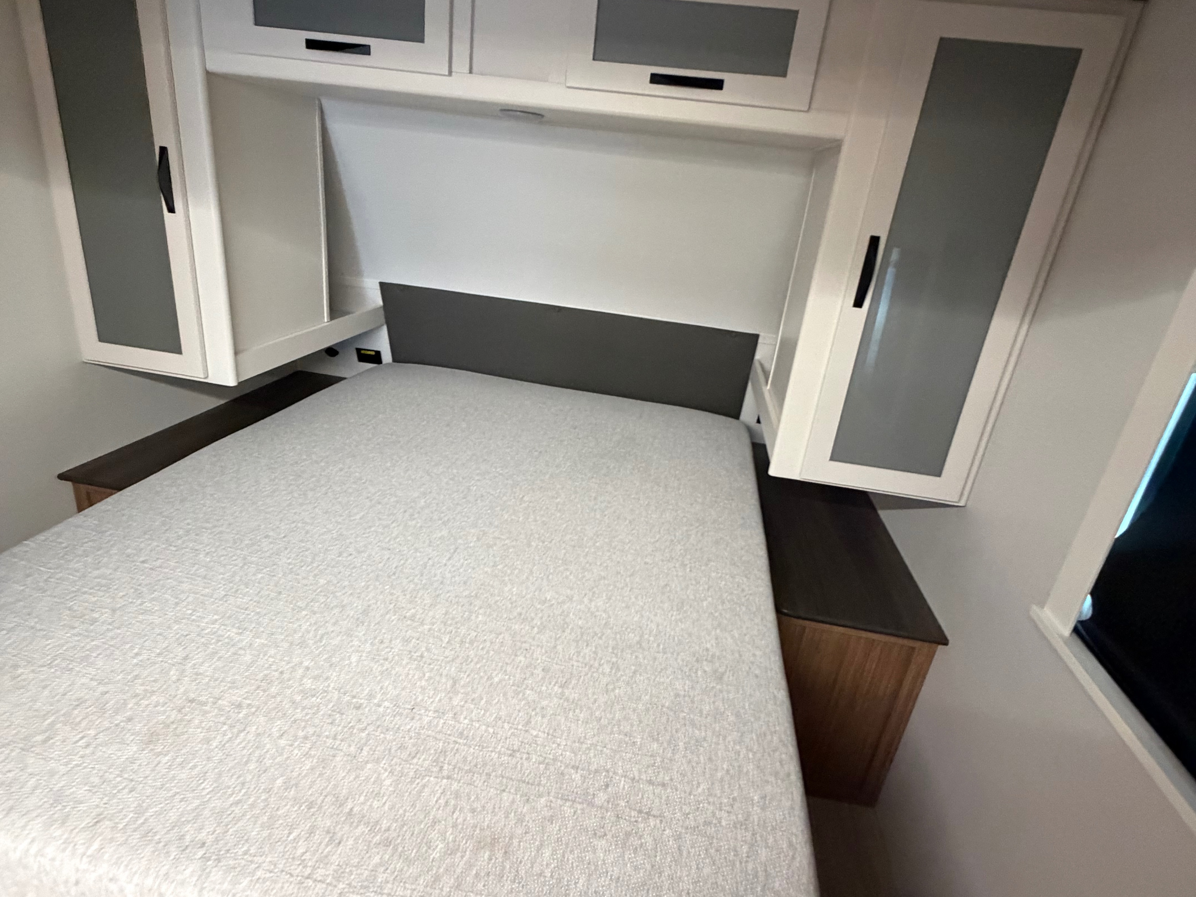 Cozy RV rental bedroom with a compact bed, dark headboard, overhead storage cabinets, and a bedside charging port.. Keystone RV Passport Super Lite 2025