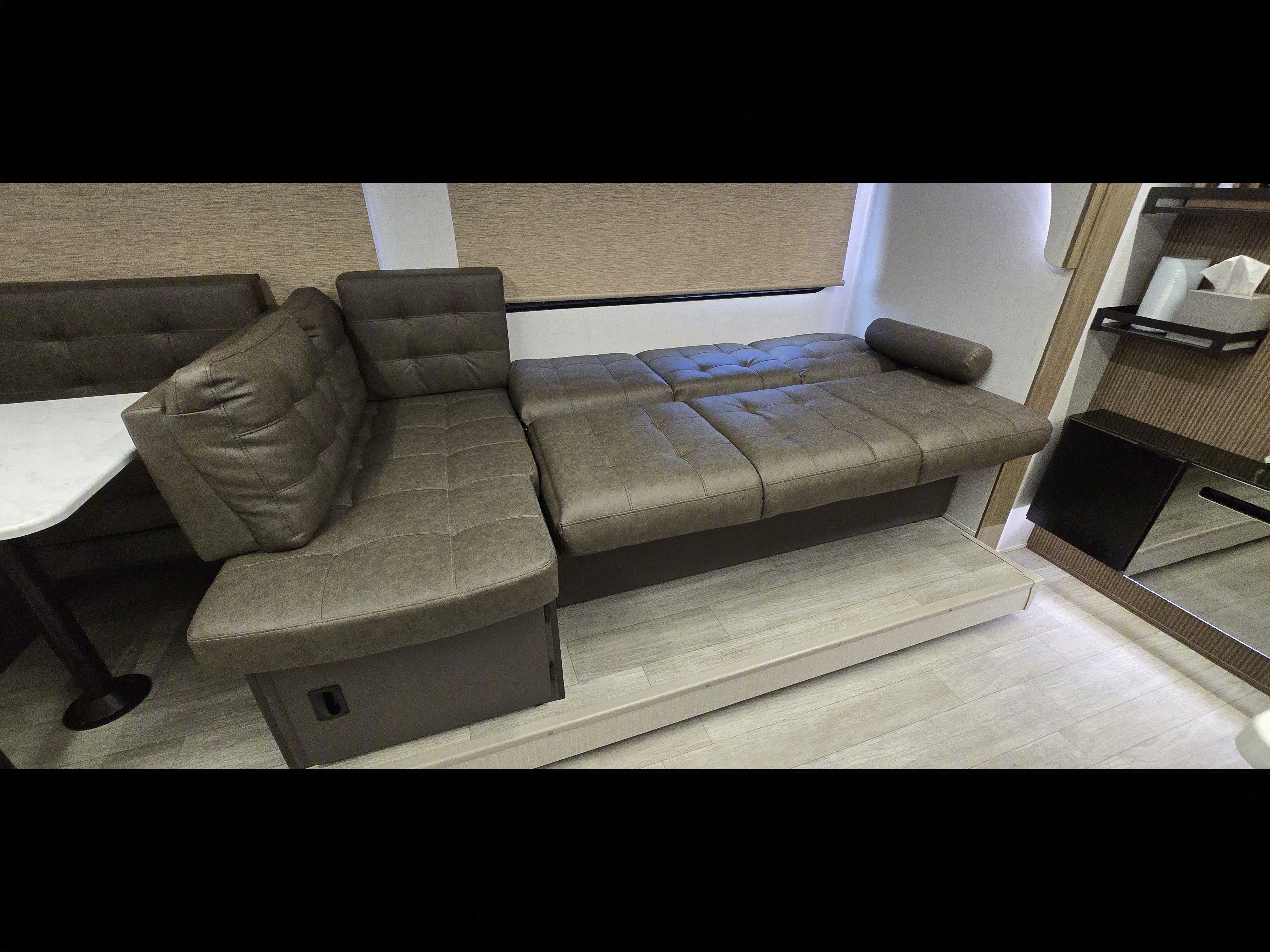 Modern RV rental interior with a brown L-shaped sofa, convertible bed extension, a small dinette, and compact storage.. Forest River Wildwood X-Lite 2025