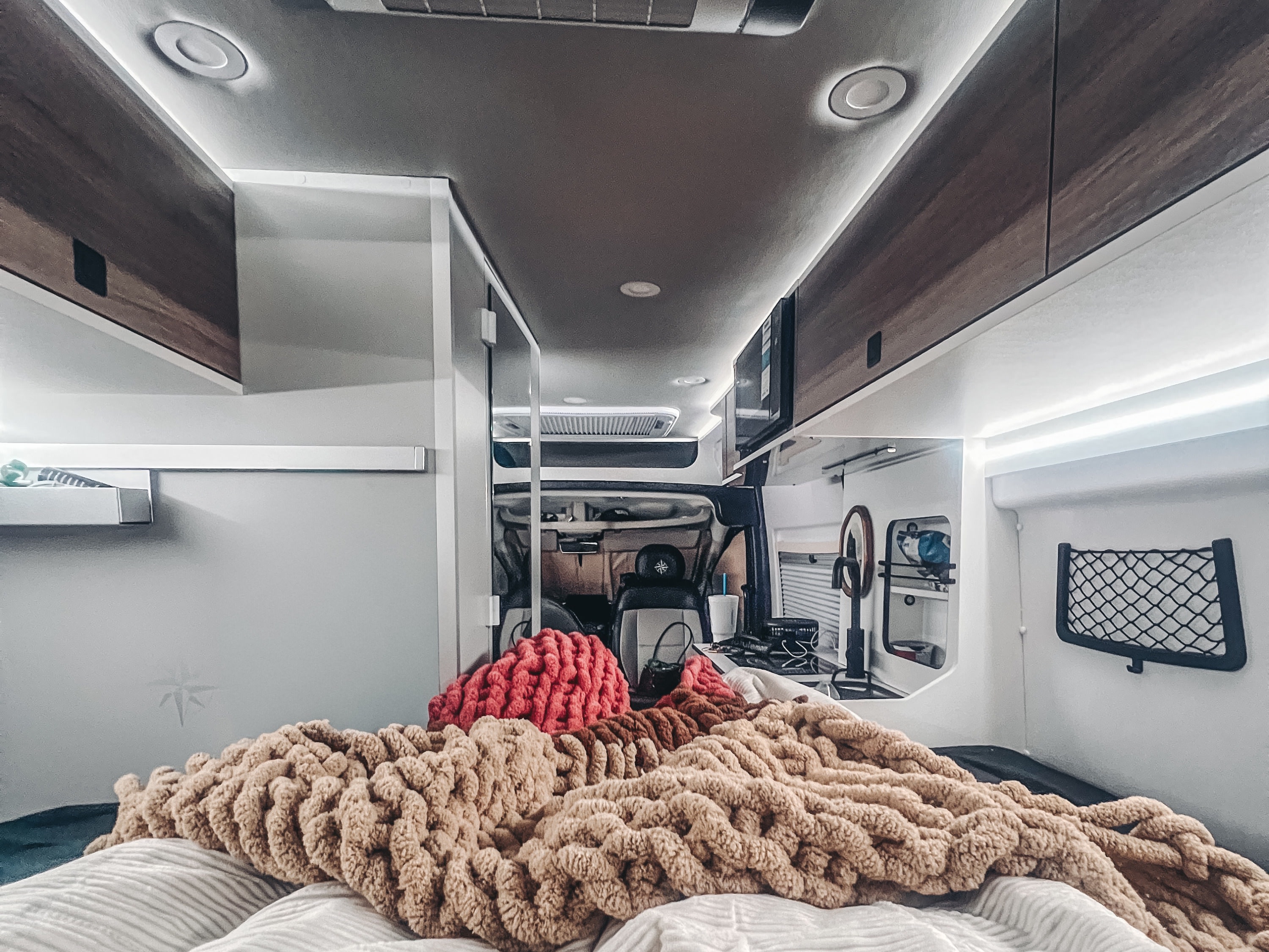 Cozy RV rental interior from the bed, featuring chunky blankets, overhead lights, and a view to the cockpit/kitchen.. Roadtrek Westfalia Wave SRT 2026