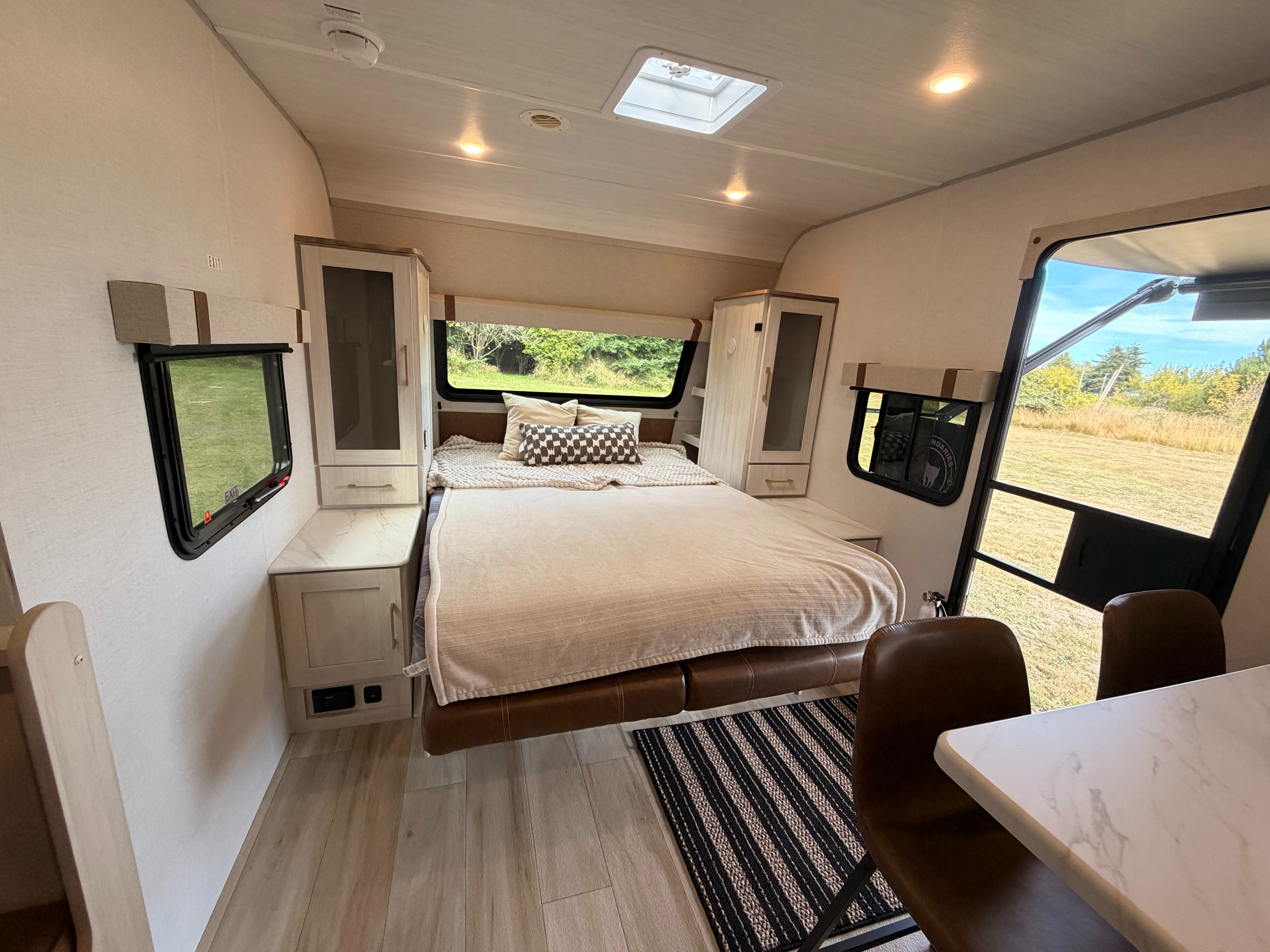 Bright RV rental interior: bedroom with bed, side cabinets, dining table with chairs, and scenic windows revealing nature.. No Boundaries 20 2026
