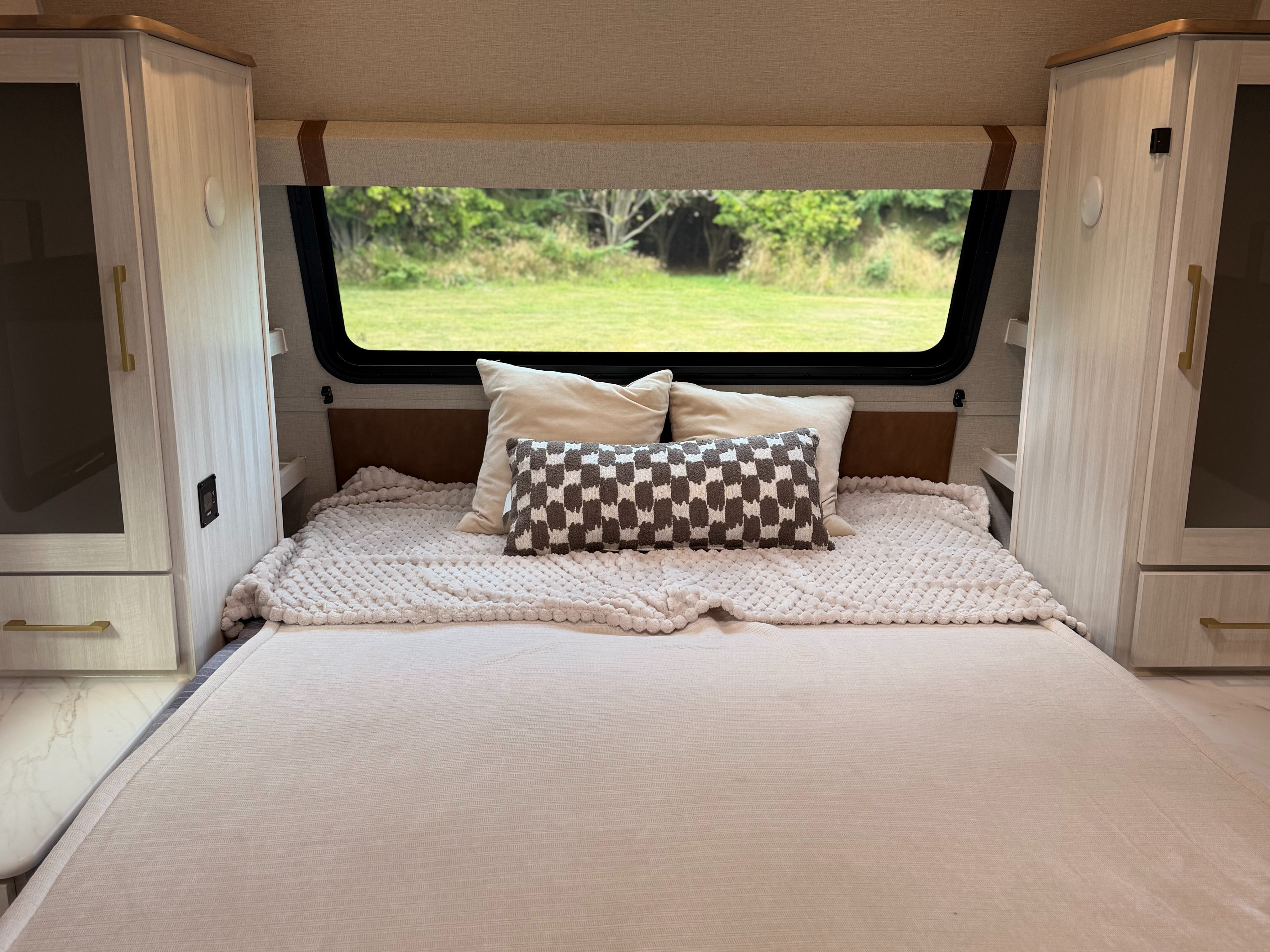 Cozy RV rental bedroom featuring a comfortable bed, light bedding, storage cabinets, and a scenic window view of nature.. No Boundaries 20 2026