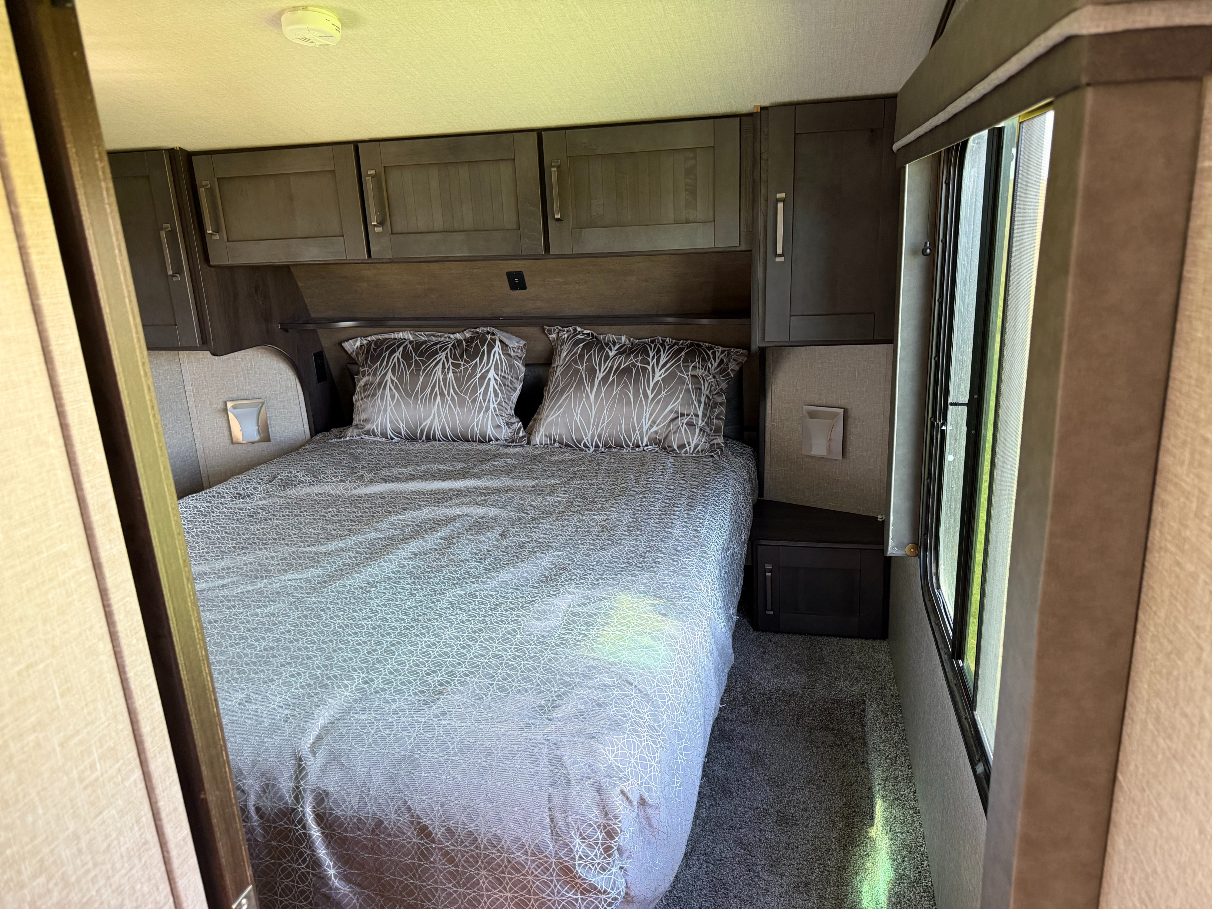 Cozy RV rental bedroom with a patterned bed, overhead cabinets, and a window, offering comfortable travel accommodation.. Grand Design Reflection 2022