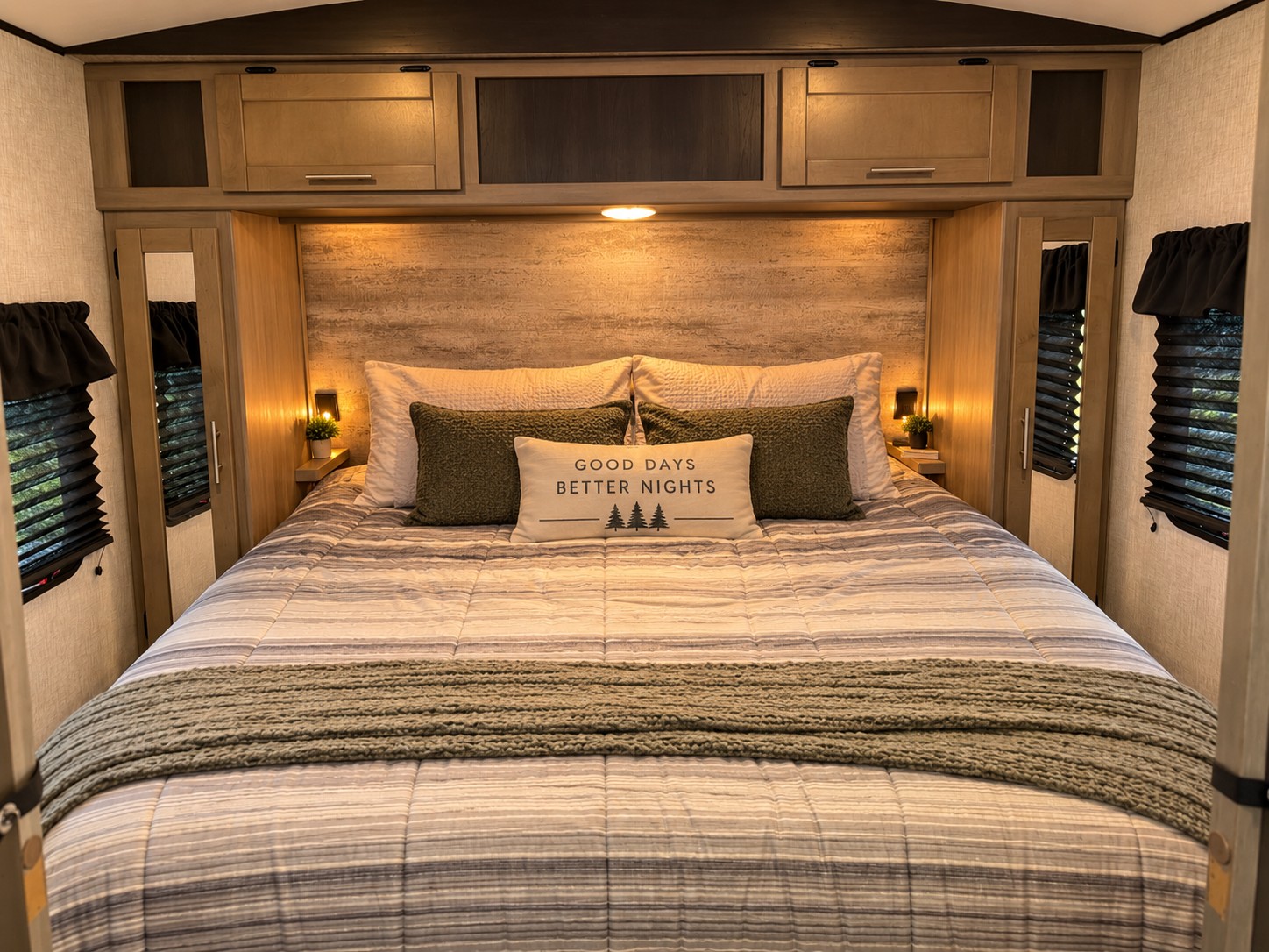 Relax in this cozy RV rental bedroom. It features warm lighting, wood accents, striped bedding, and overhead cabinets for storage.. Crossroads RV Sunset Trail Super Lite 2022