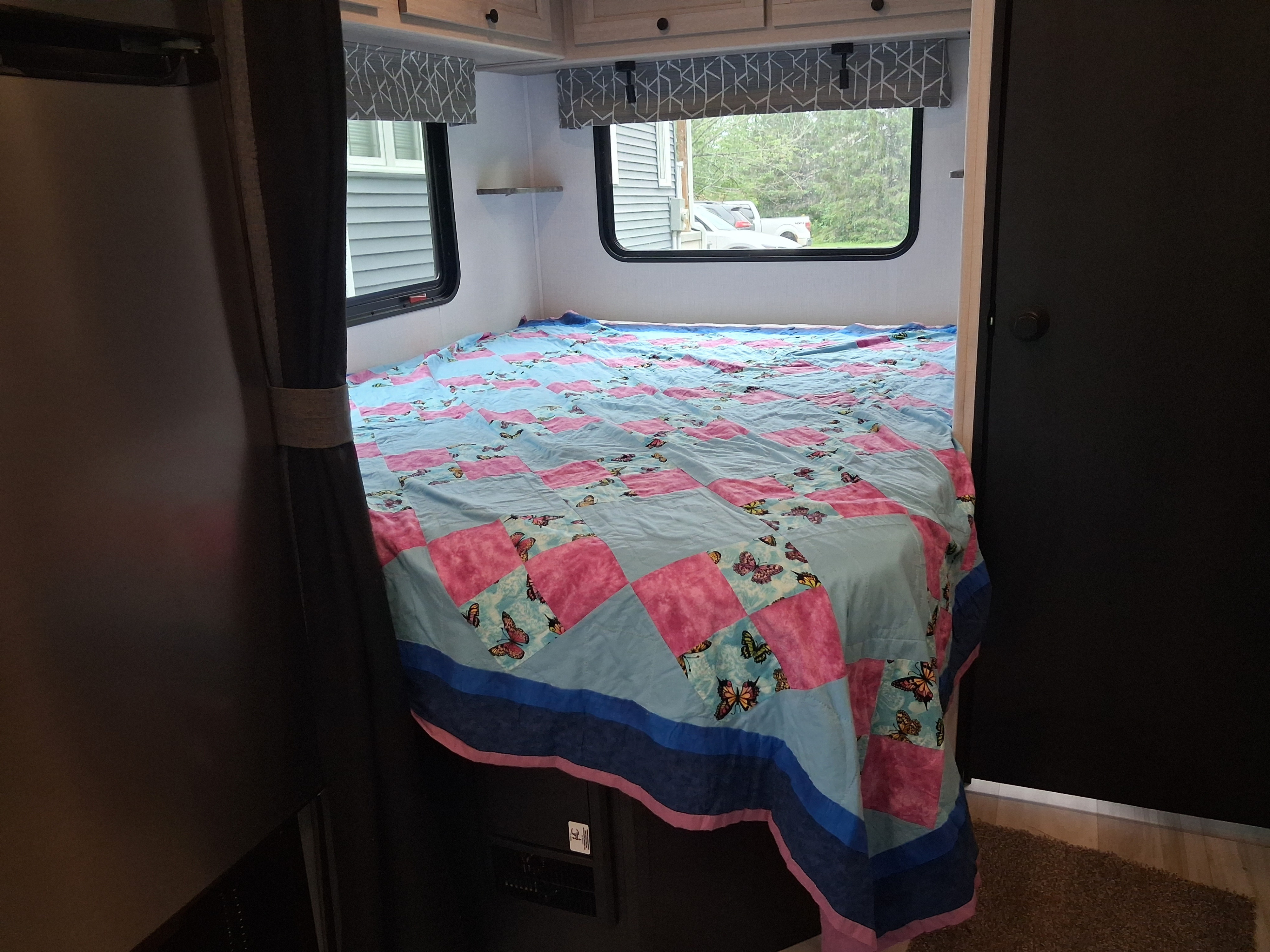 Cozy RV rental bedroom with a blue and pink butterfly quilt on the bed, cabinets, and windows looking out to nature.. FOREST RIVER SL 2150LE 2027