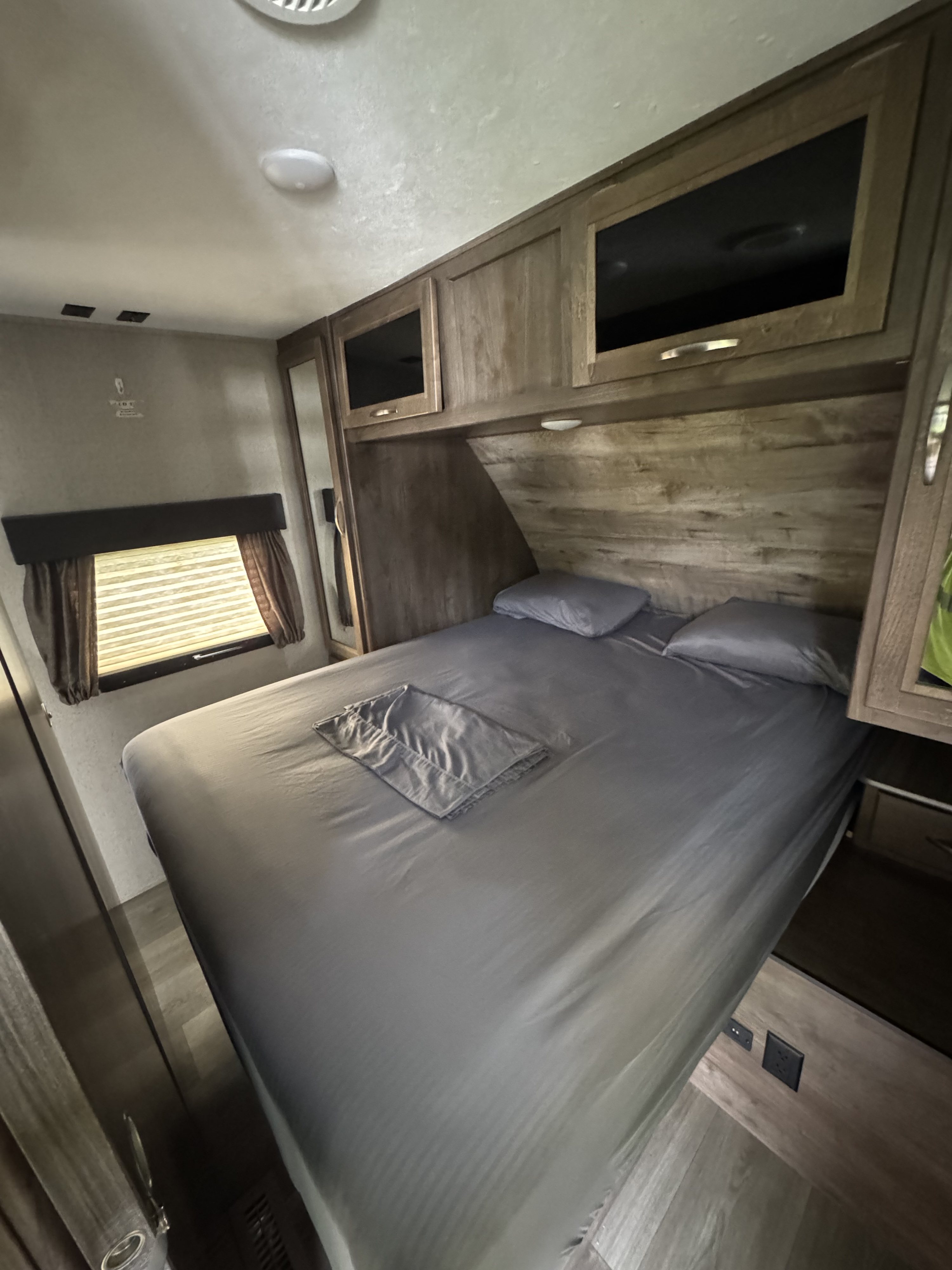 Inside an RV rental, this bedroom features a comfy grey bed, wood cabinets, and a window for natural light.. Dutchmen Coleman Light 2019