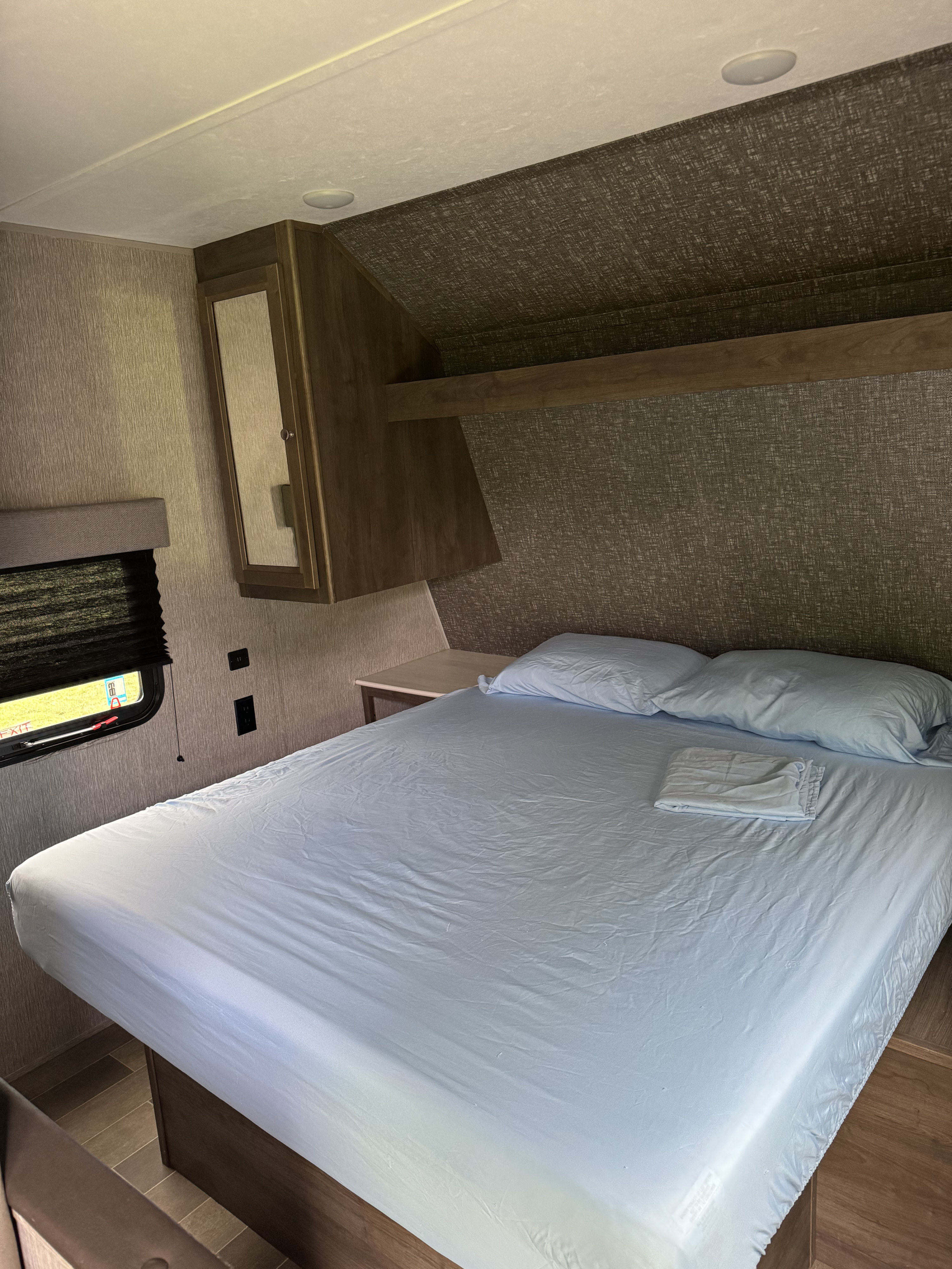 A compact rv rental bedroom showcasing a bed with light blue sheets, pillows, a window, and wood cabinetry.. Dutchmen Coleman Lantern LT 2020