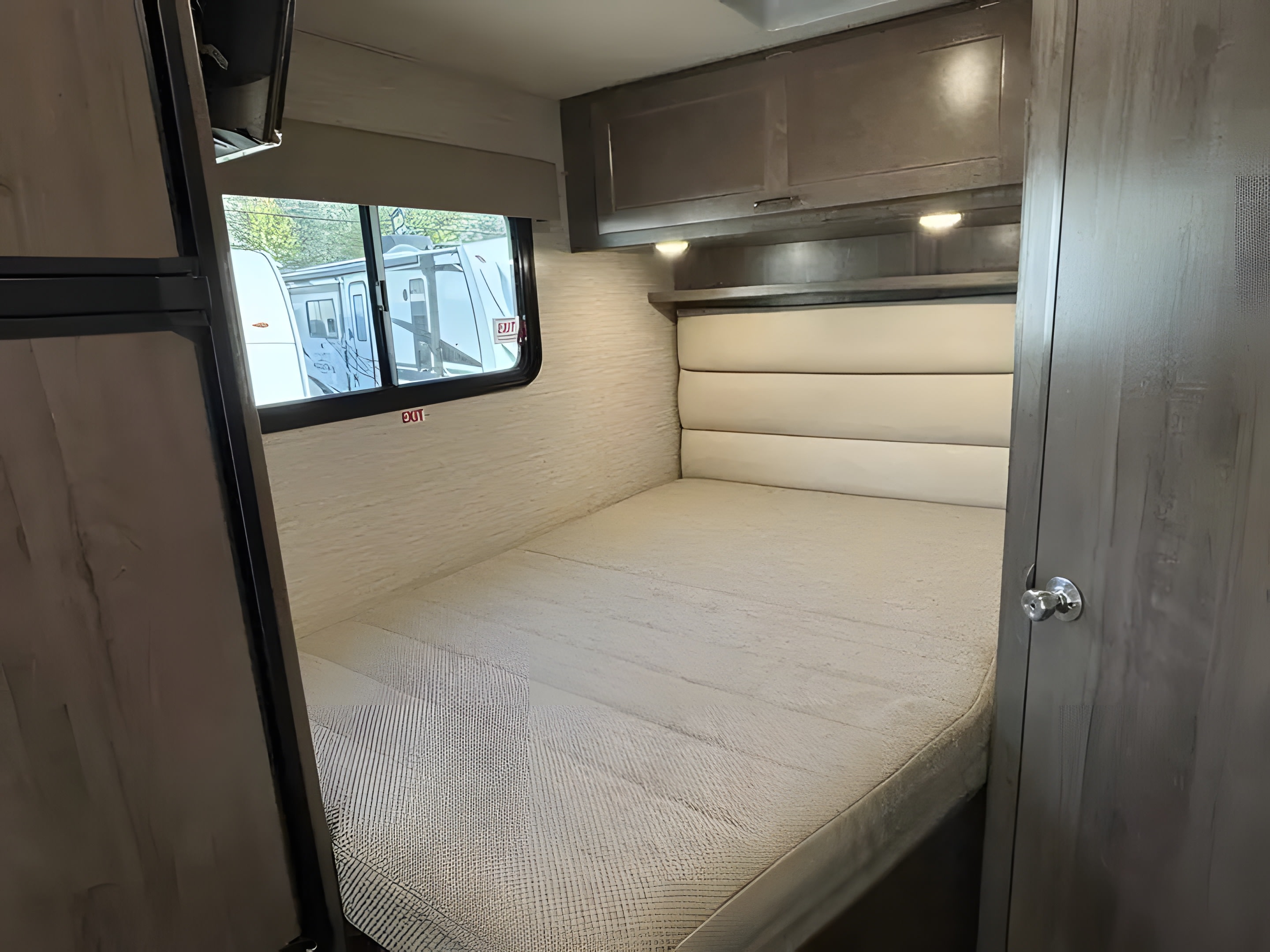 Cozy RV rental bedroom with a bed, padded headboard, overhead cabinets, and a window showing another RV outside.. Winnebago Minnie Winnie 2022