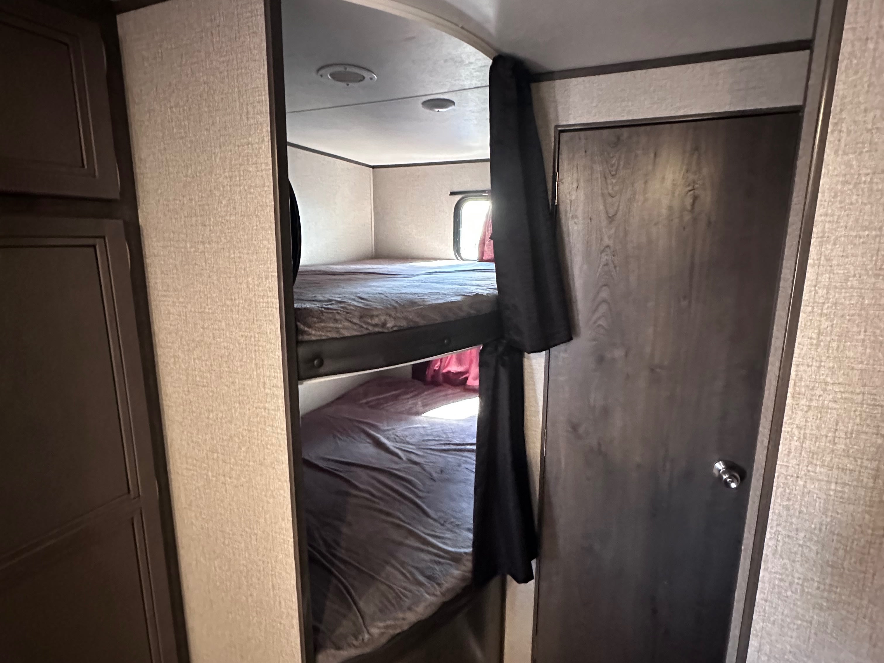 Inside an RV rental: two bunk beds with gray bedding, a partially drawn black curtain, and a dark wood door in a compact camper interior.. Jayco Jay Flight SLX Rocky Mountain Edition 2020