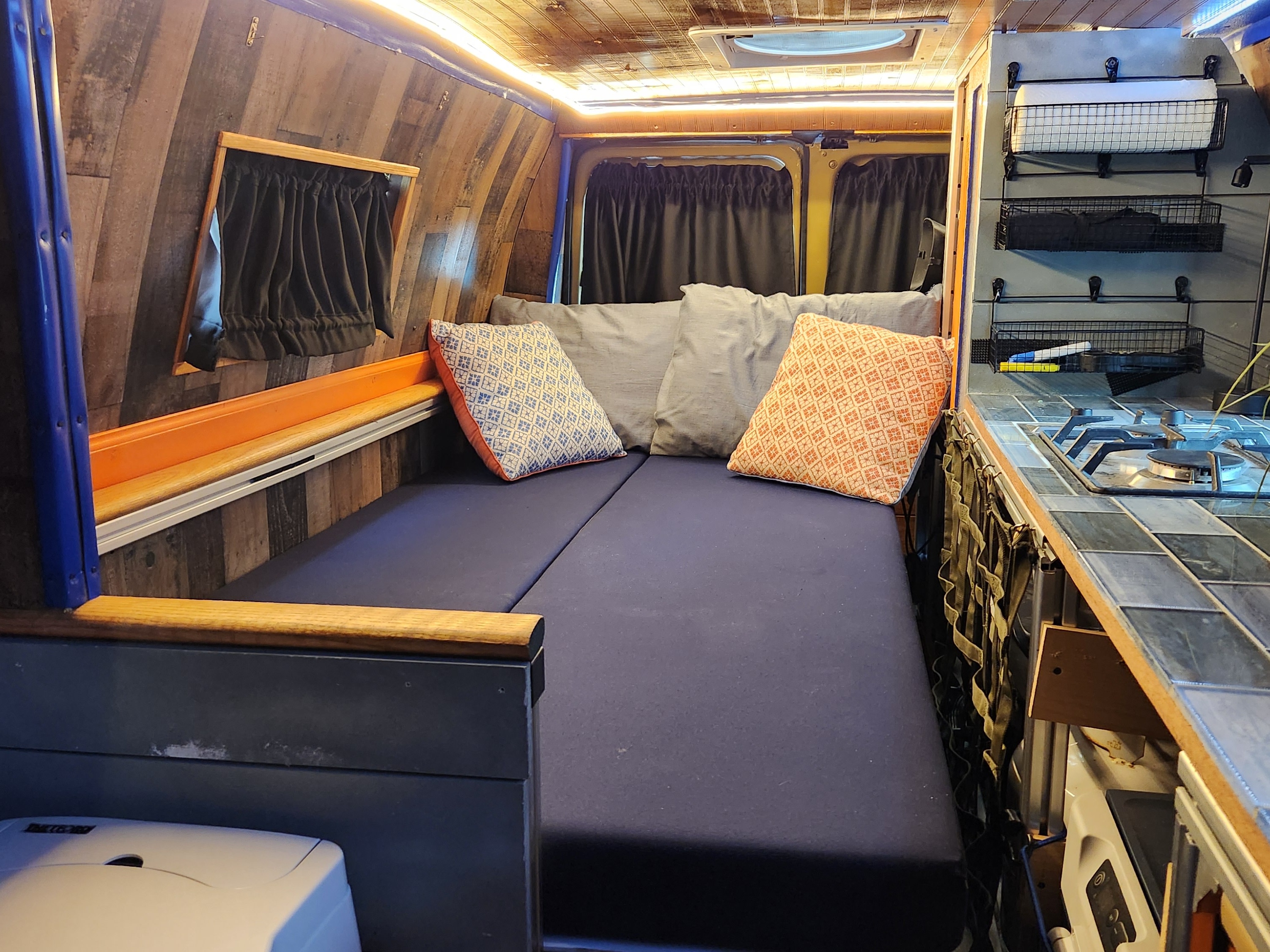 Cozy rv rental camper van interior with a bed, wood-paneled walls, LED lighting, and a compact kitchen stove area.. Ford E350 Box Van 2006
