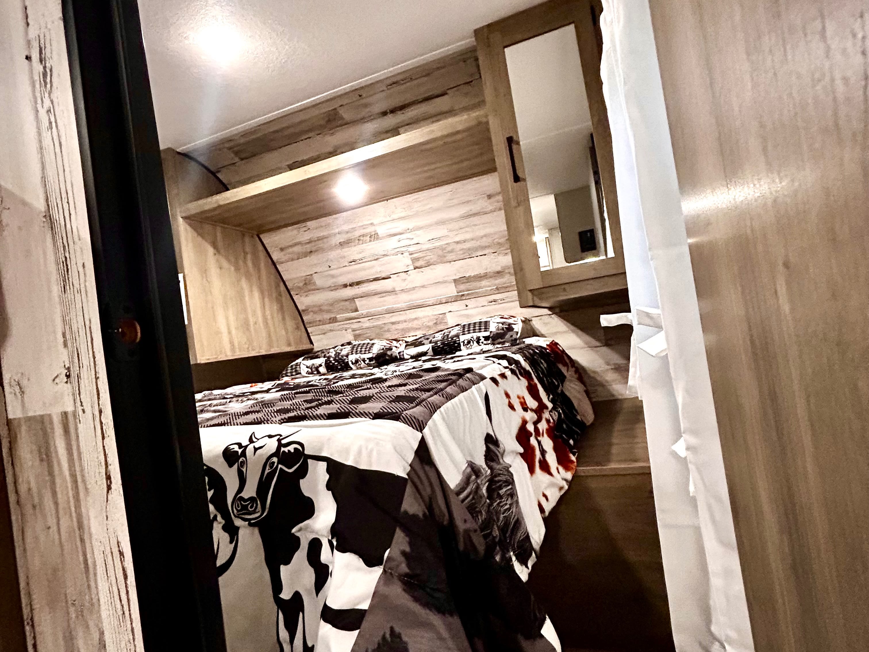 Cozy RV rental bedroom featuring a cow print comforter, light wood paneling, overhead shelf, and mirrored cabinet.. Forest River Palomino Puma XLE Lite 2021