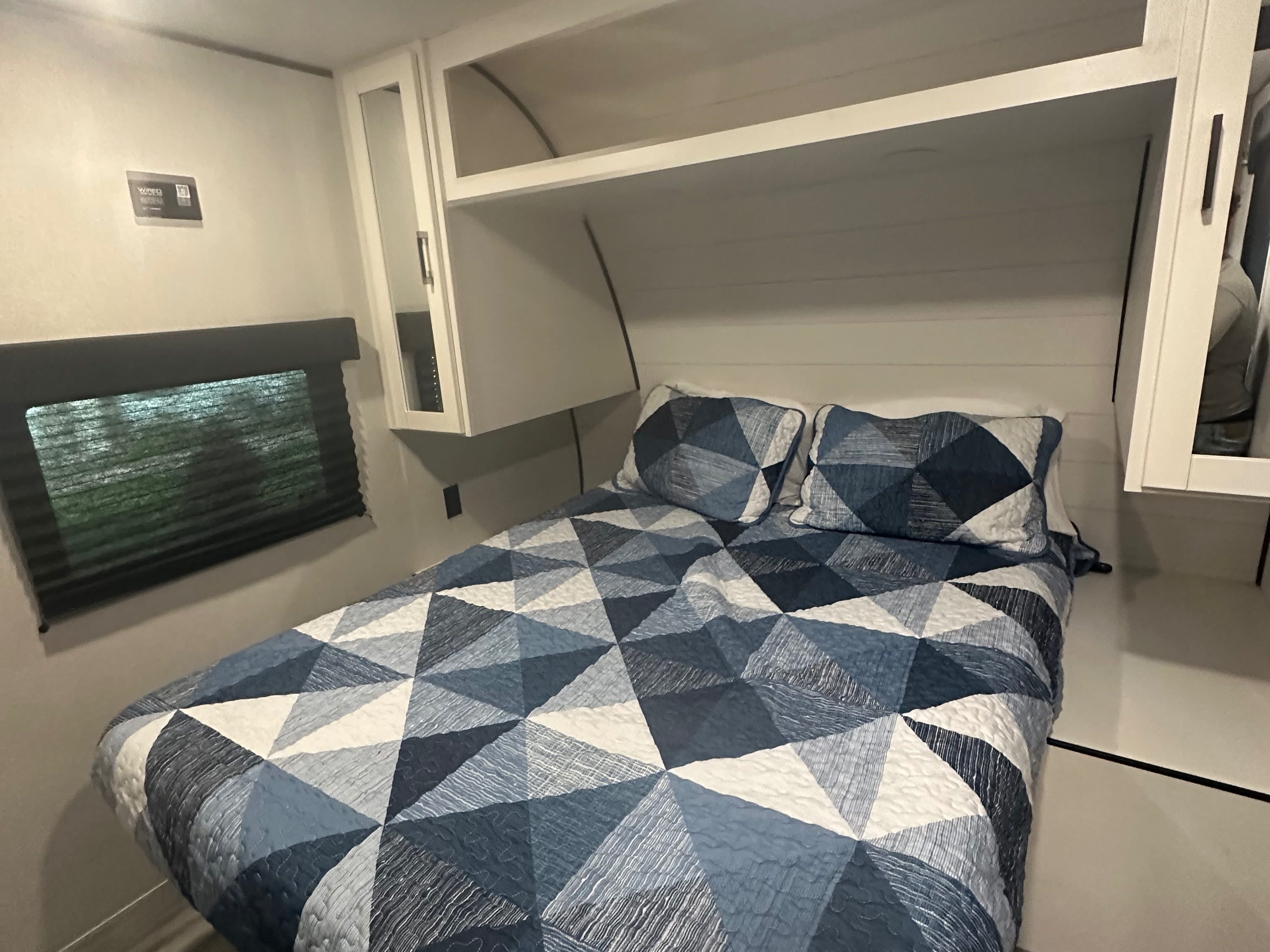 This RV rental bedroom has a comfortable bed with blue geometric patterned bedding, overhead cabinets, and a window with blinds.. Jayco Jay Flight SLX 2026