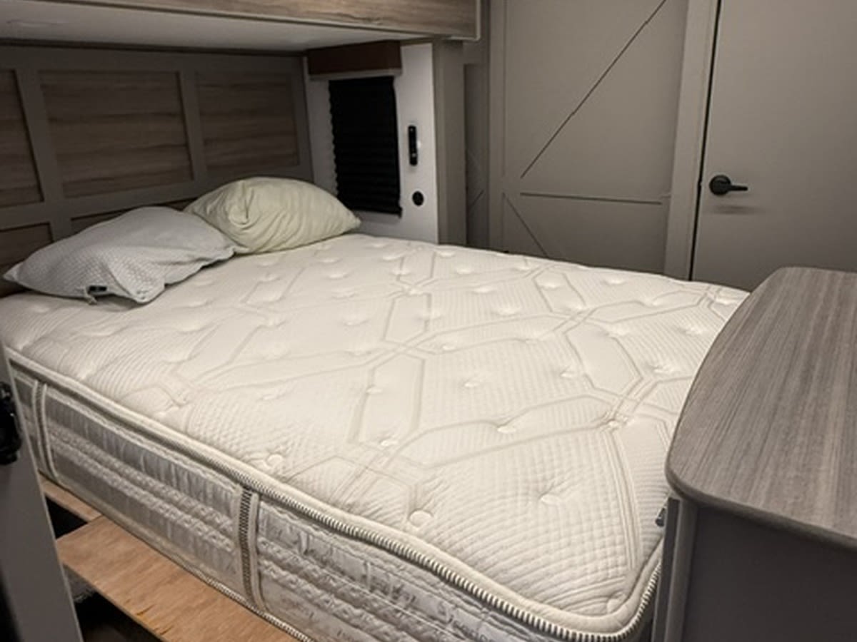 Comfortable RV rental bedroom with a white mattress, pillows, wooden headboard, and a small cabinet, offering a cozy sleep space.. Keystone RV Sprinter 2025
